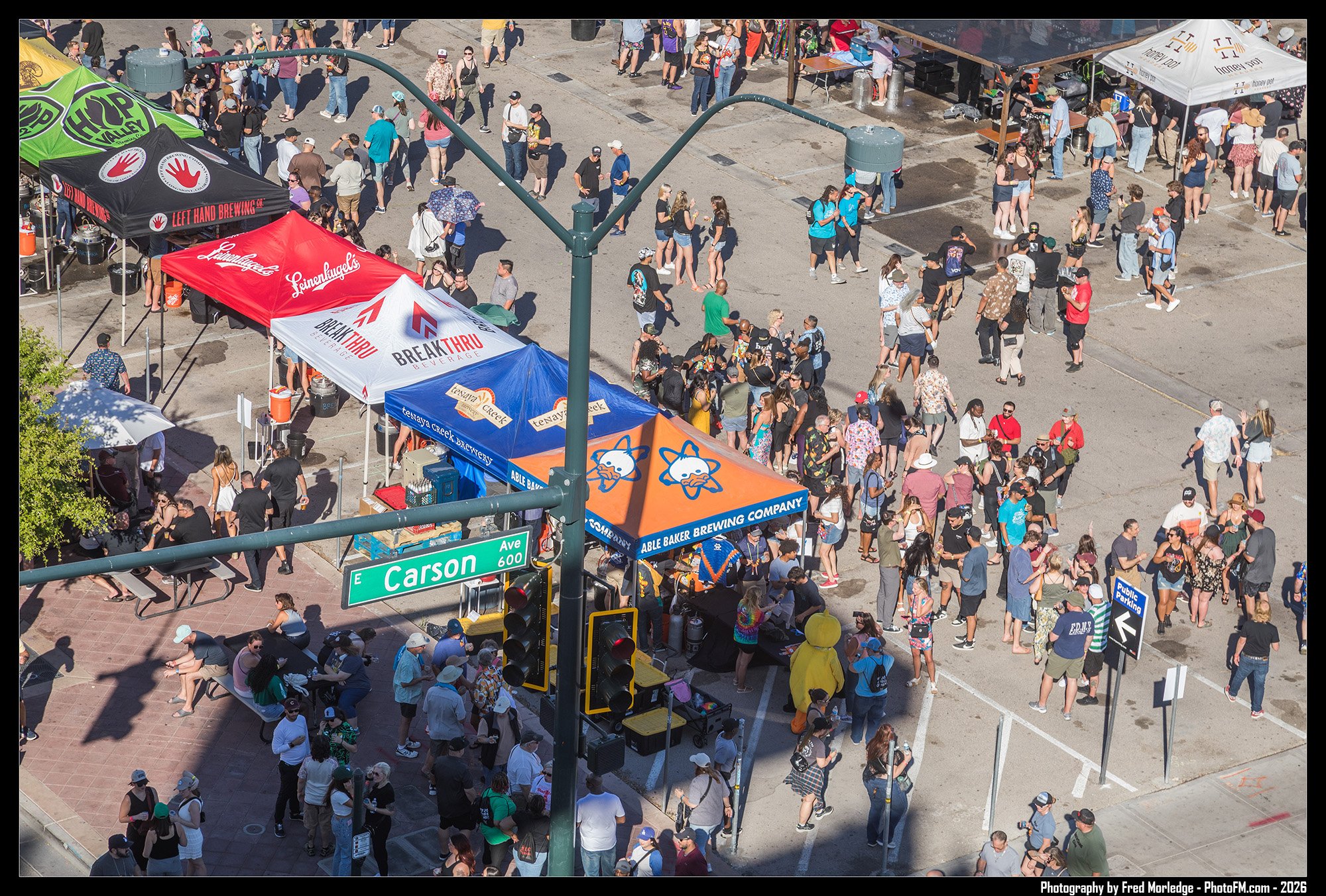Crowd of people attending outdoor event with vendor tents on a city street corner at Carson Avenue, including tents for local breweries and food vendors, with some people sitting and others walking around.