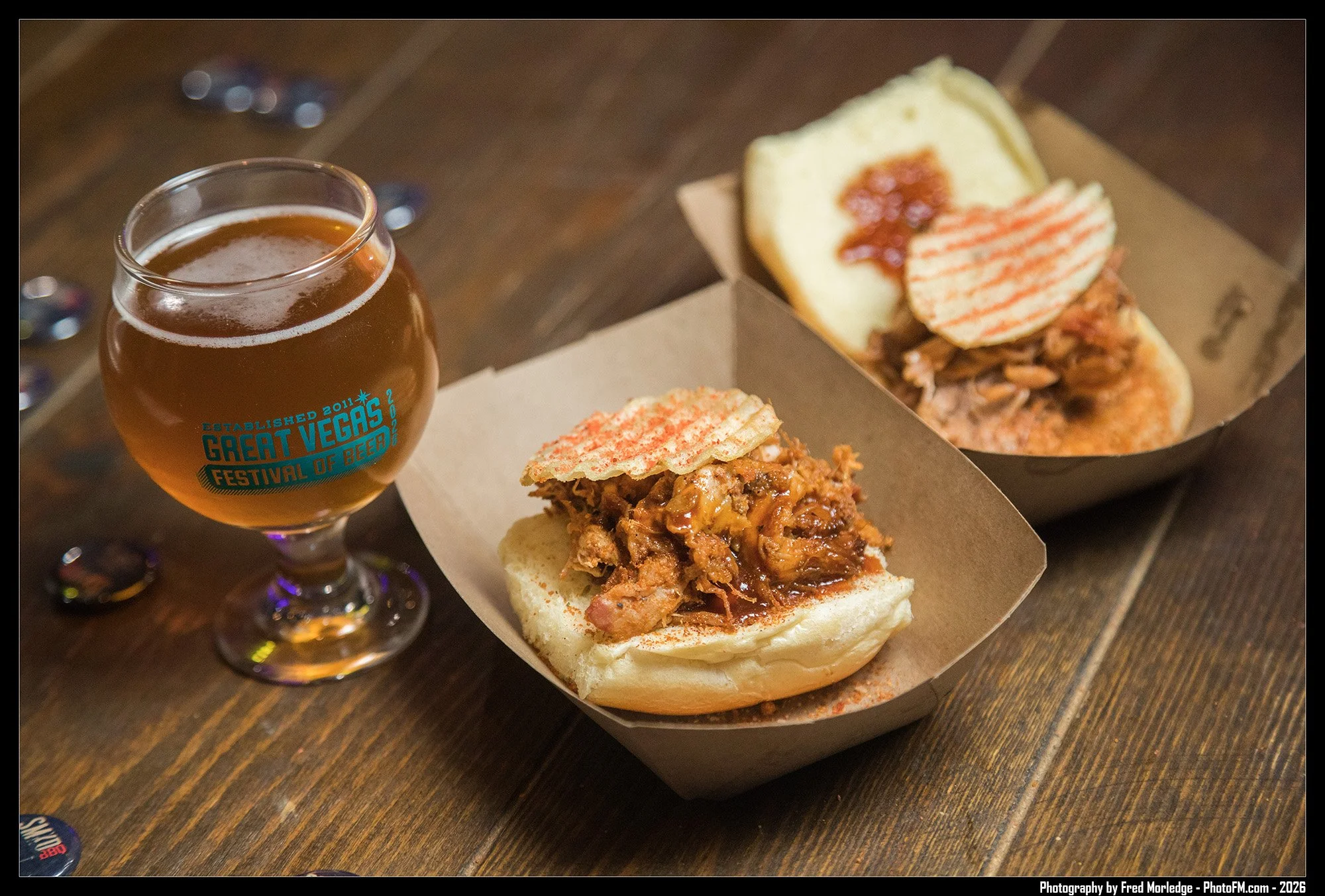 A glass of beer next to two barbecue sandwiches on a wooden table.