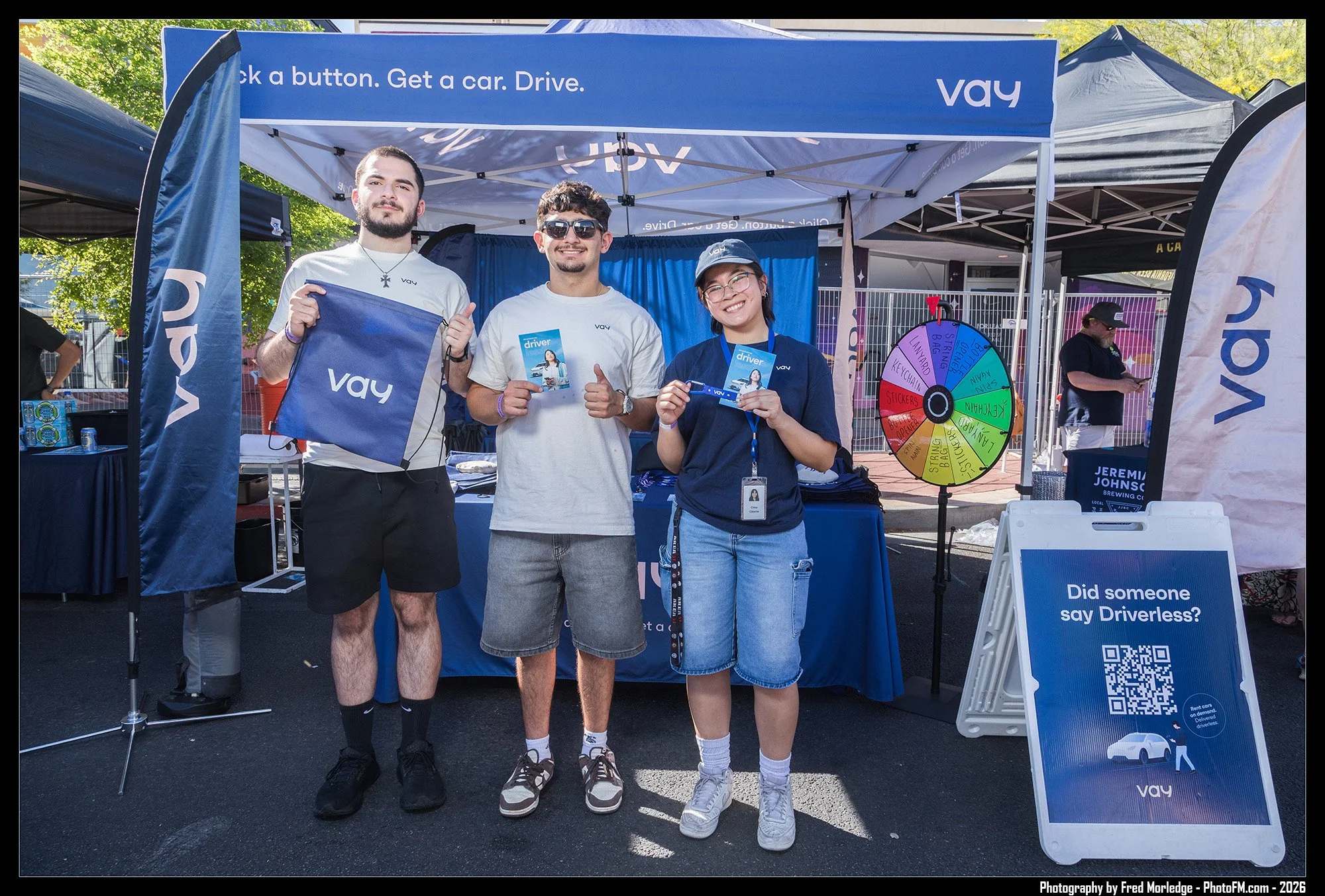 Three young people standing in front of a blue promotional booth with a spinning wheel, holding promotional materials and smiling. There is a sign asking if someone said 'Driverless?' and a QR code on a stand.