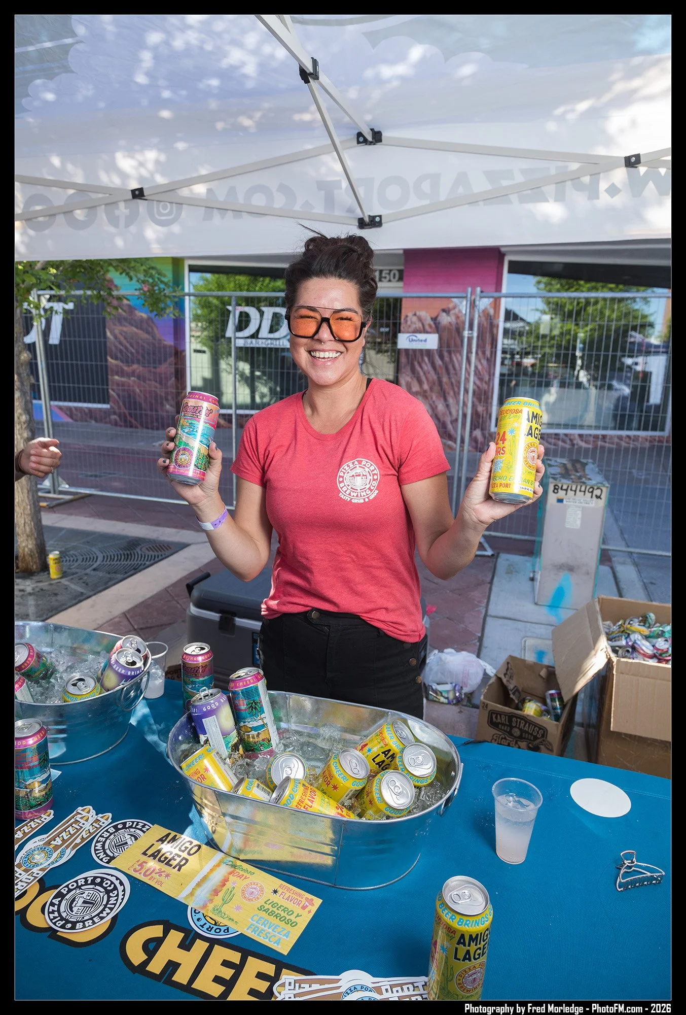 Woman smiling, holding colorful cans of lager beer at an outdoor event booth with Pepsi cans and promotional materials on the table.