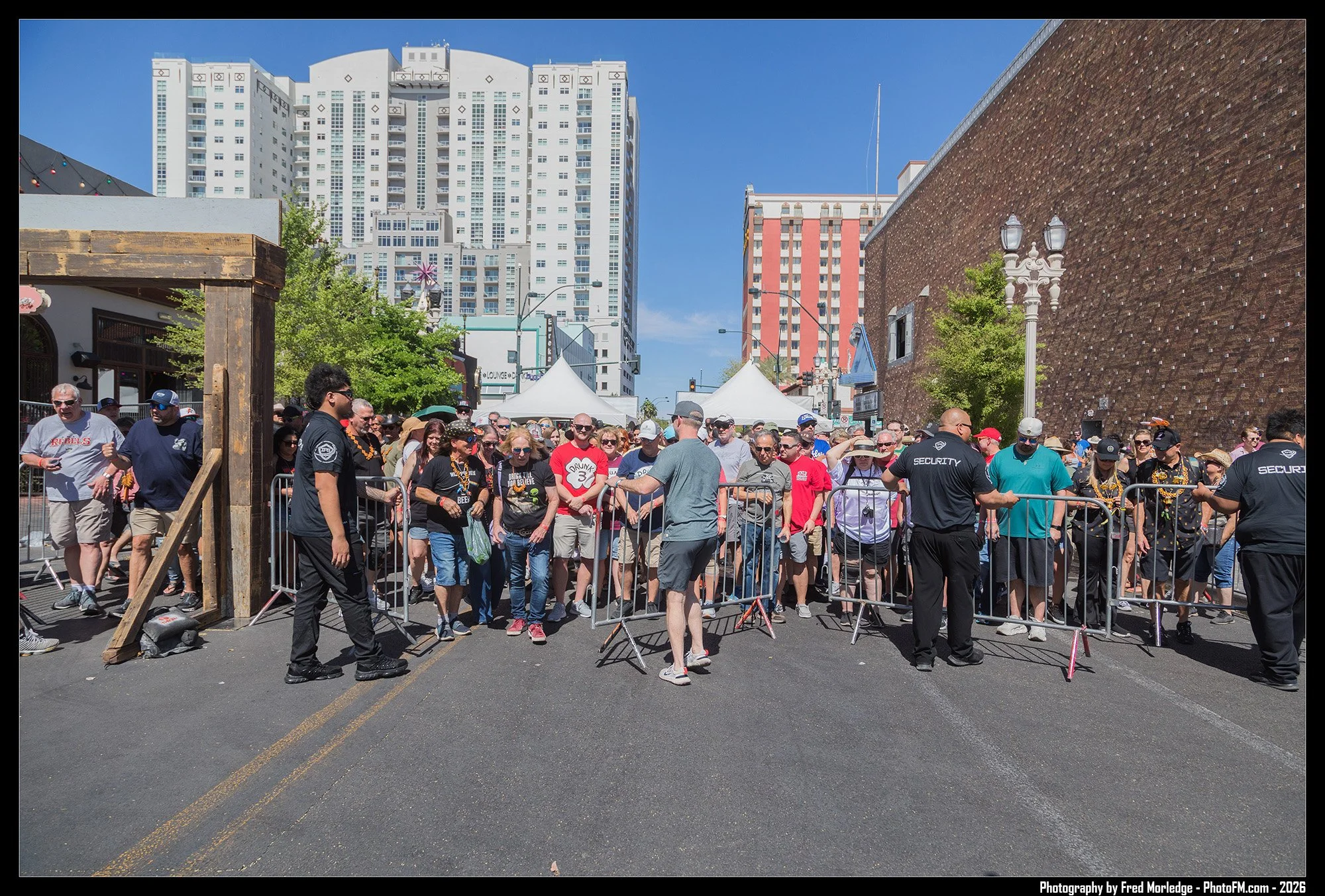 Crowd of people waiting behind metal barricades on an urban street during a sunny day, with high-rise buildings and tents in the background.