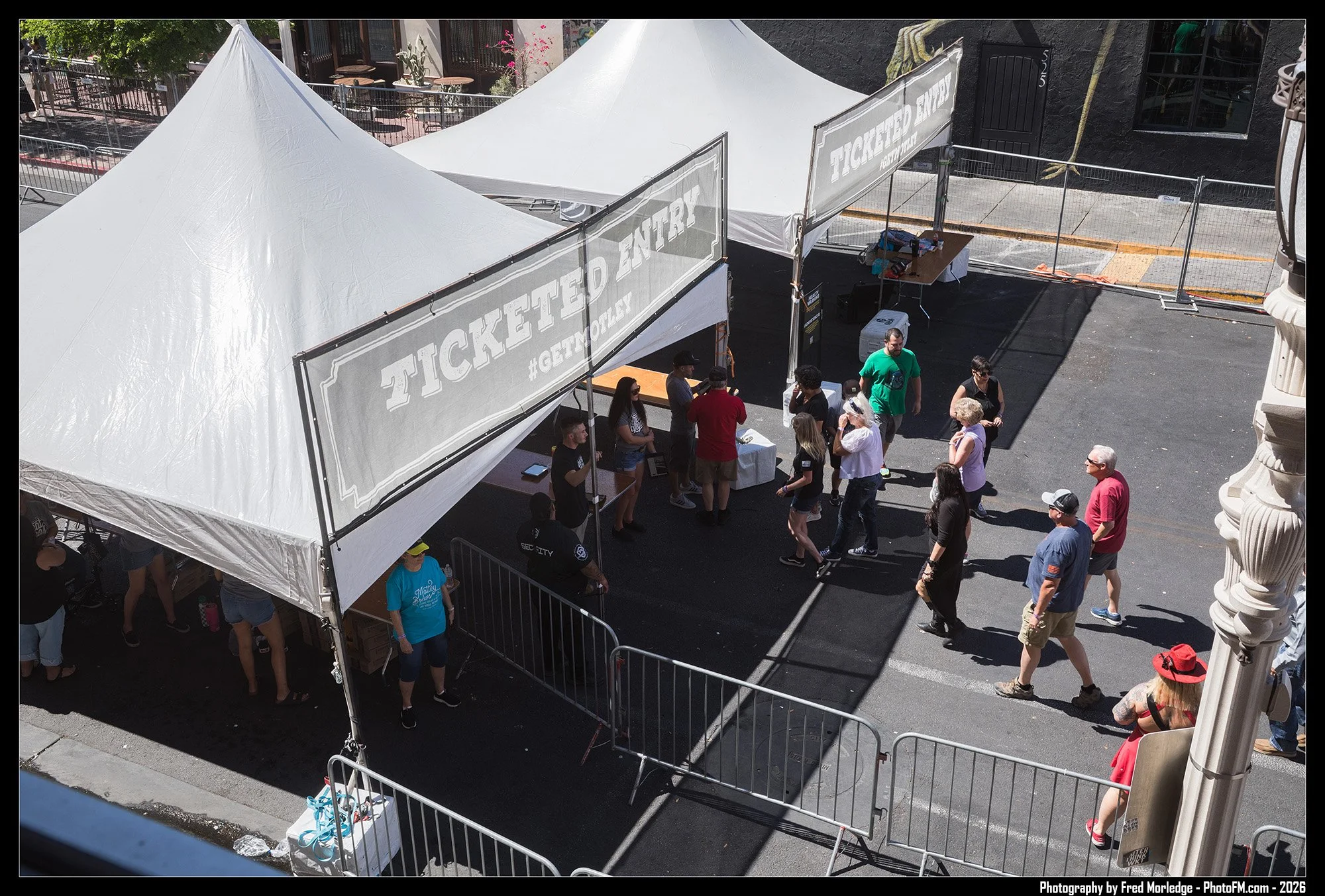 An outdoor scene with white tents labeled 'Ticketed Entry' and a crowd of people walking and standing around. There is a metal fence and a sidewalk with a black building in the background.