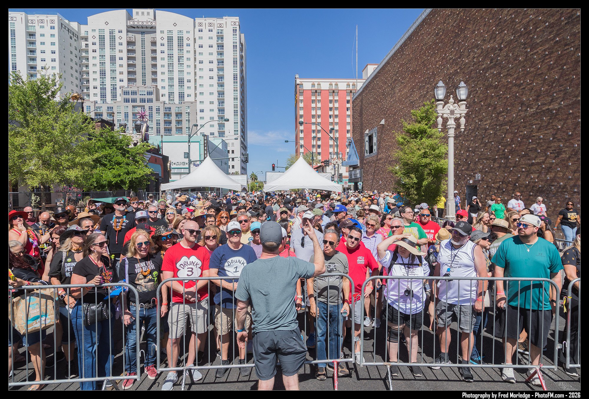 A large crowd of people standing behind metal barricades at an outdoor event on a sunny day with tall buildings and tents in the background.