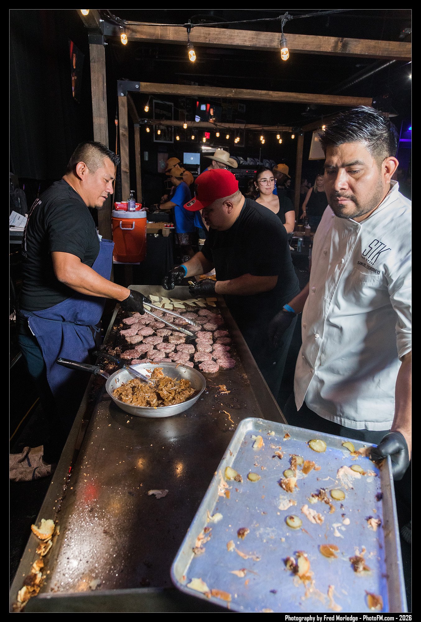 Three men are preparing and cooking hamburger patties and sliced onions on a large grilling surface in a dimly lit indoor space. One man is dressed as a chef, observing the process, while the others are actively grilling meat.