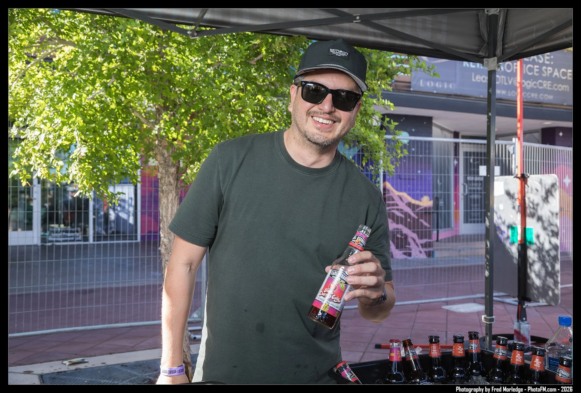 A smiling man wearing sunglasses and a black cap holding a bottle of cherry soda at an outdoor event with a booth displaying multiple bottles of soda and trees in the background.