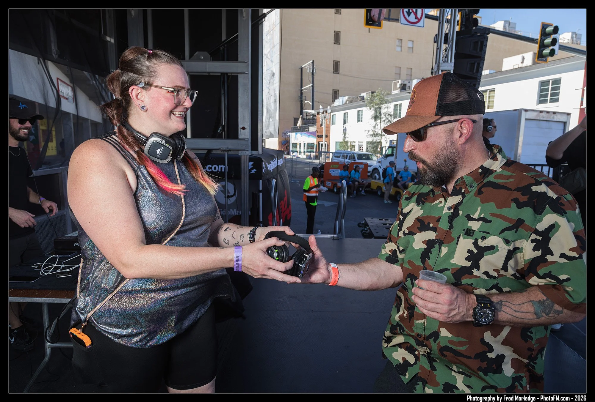 A woman with glasses and colorful hair receiving a handshake from a man in camouflage shirt and cap at an outdoor event.