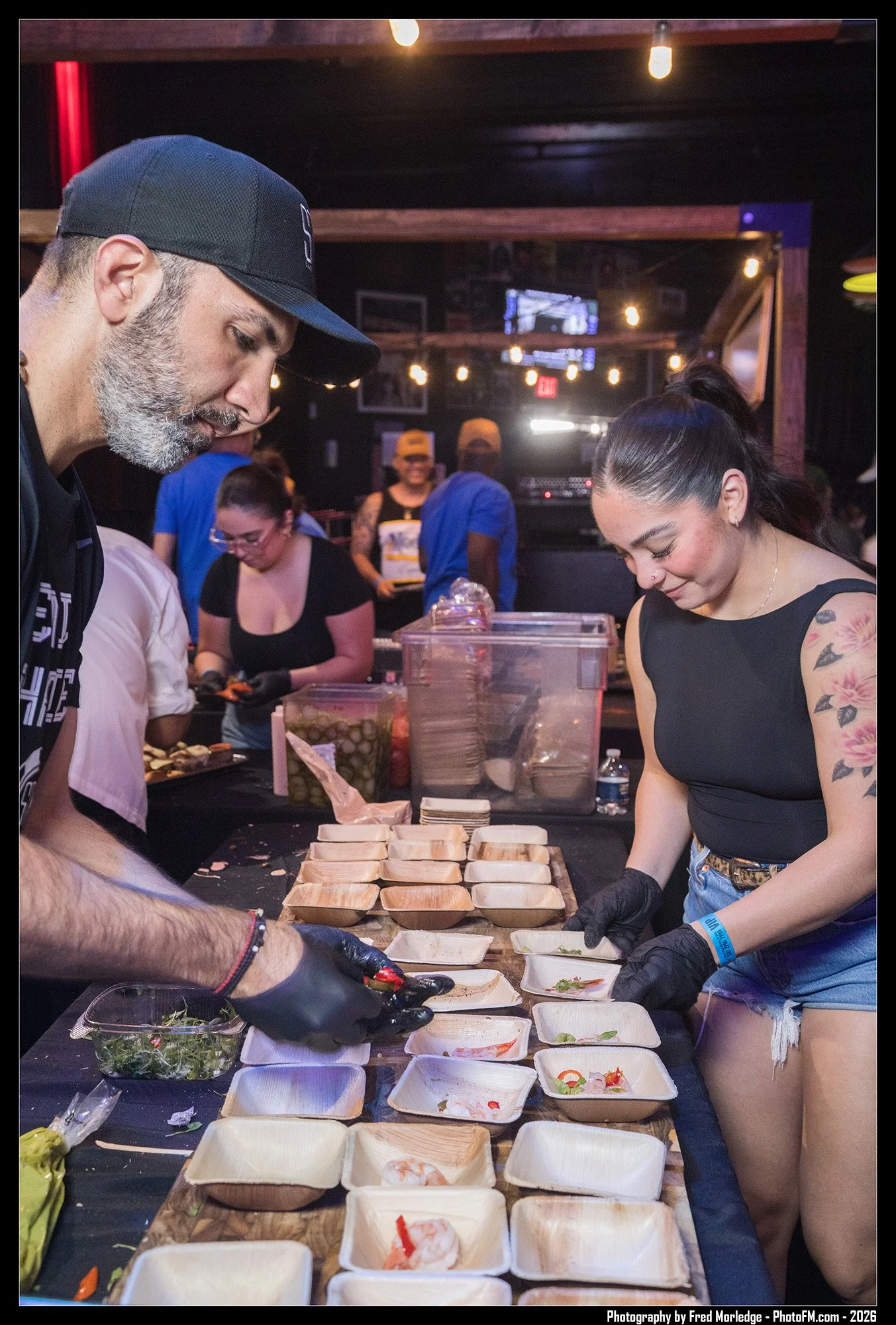 People preparing sushi at a table in a dimly lit room, with ingredients and trays of sushi. Two individuals in the foreground are focused on assembling sushi, with others working in the background.