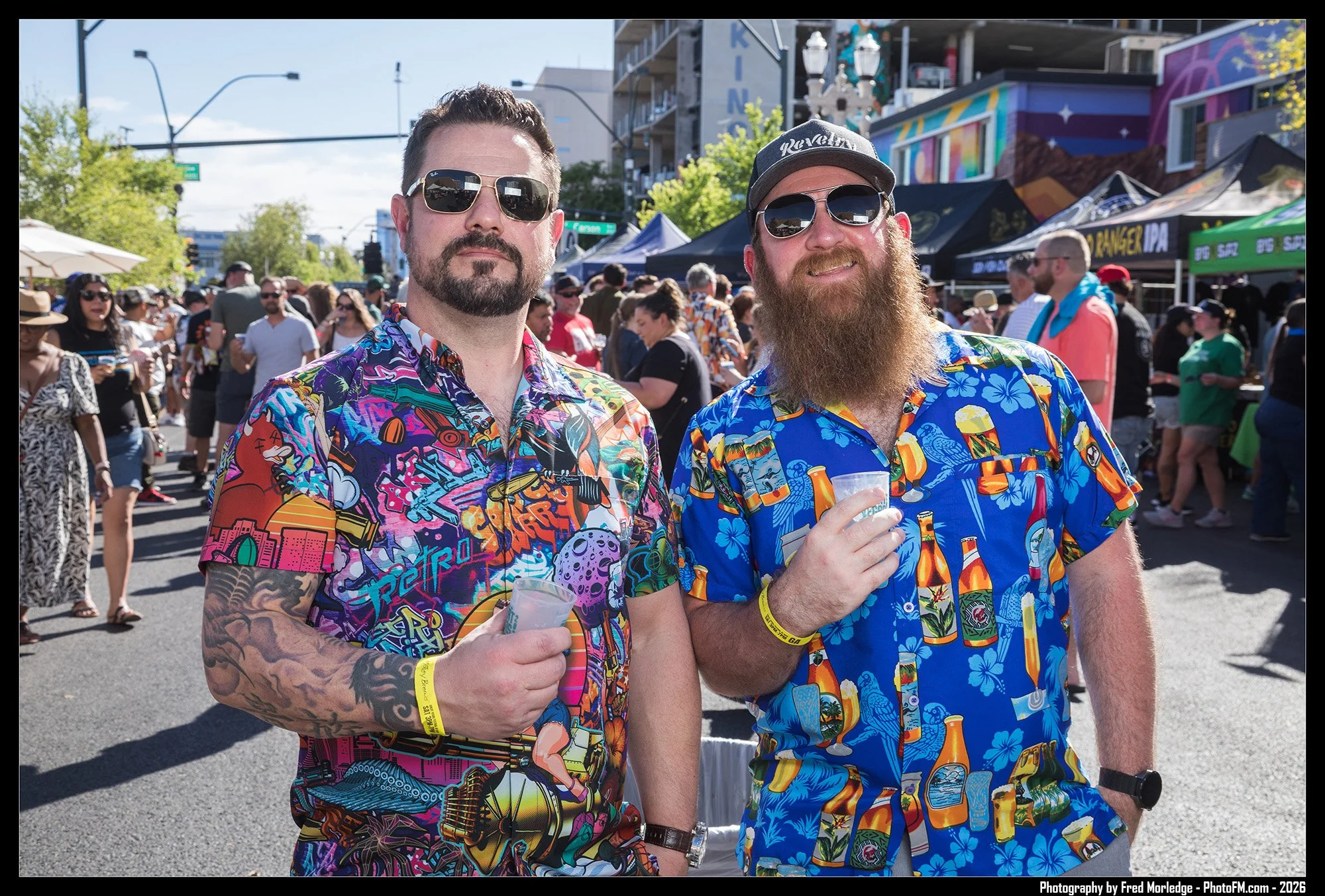 Two men wearing colorful Hawaiian shirts and sunglasses at an outdoor event with a crowd, vendor tents, and urban buildings in the background.