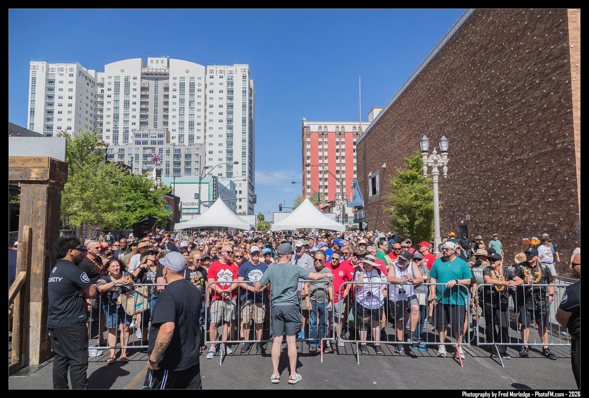 Crowd of people gathered outdoors behind a metal barricade during daytime, with tall buildings and a blue sky in the background.