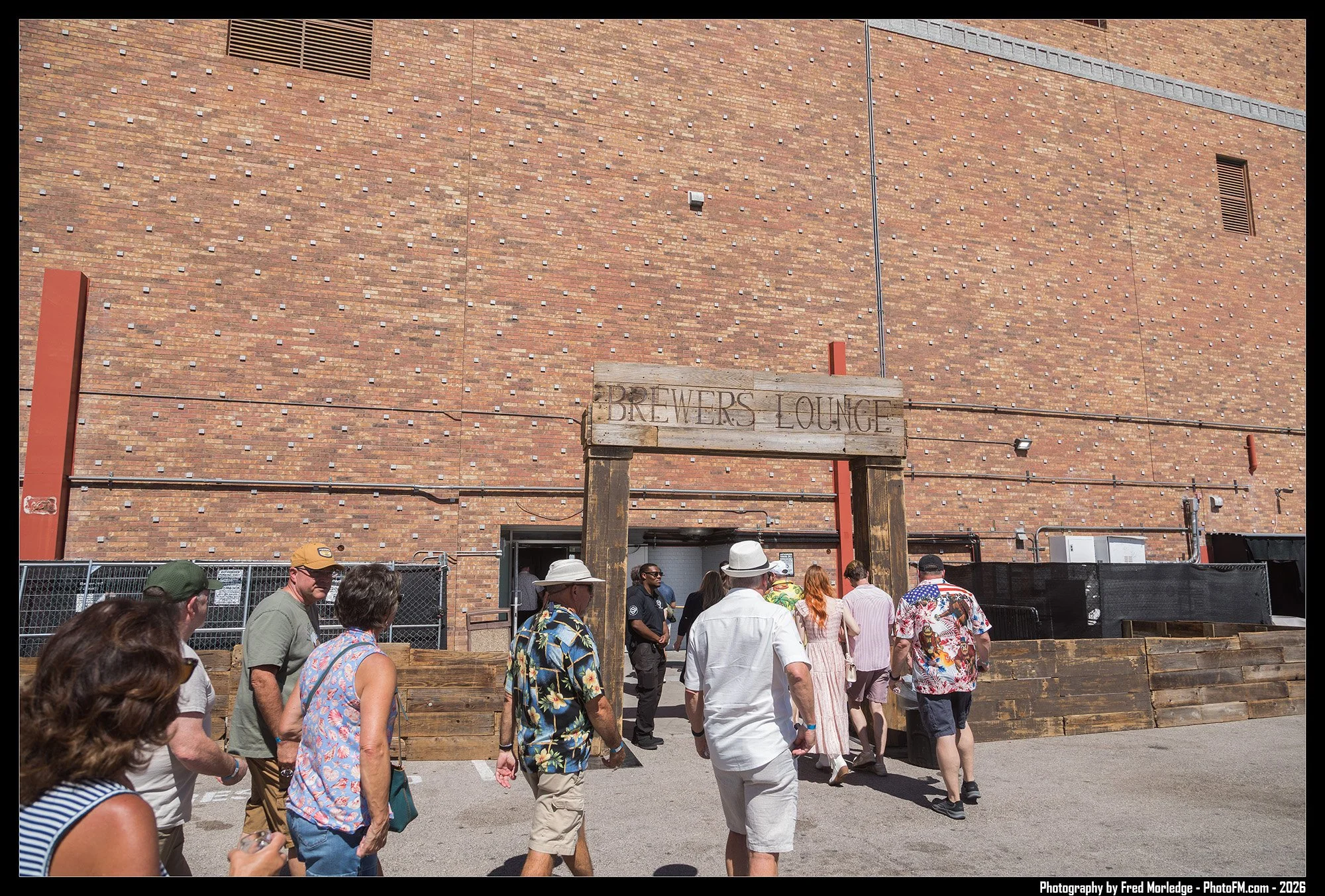 People gathering outdoors near a brick building with a wooden sign that reads 'BREWERS LOUNGE.' There is a crowd of people walking and standing, some wearing hats and casual summer clothing, in front of a fenced area with wood railings.