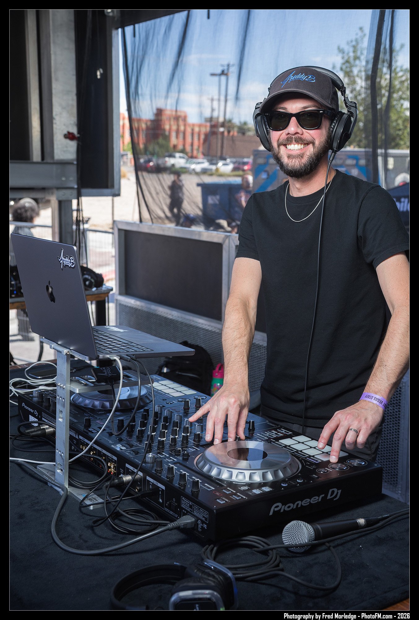 A smiling male DJ wearing sunglasses and a black baseball cap, standing behind a DJ mixer and a laptop, performing at an outdoor event.