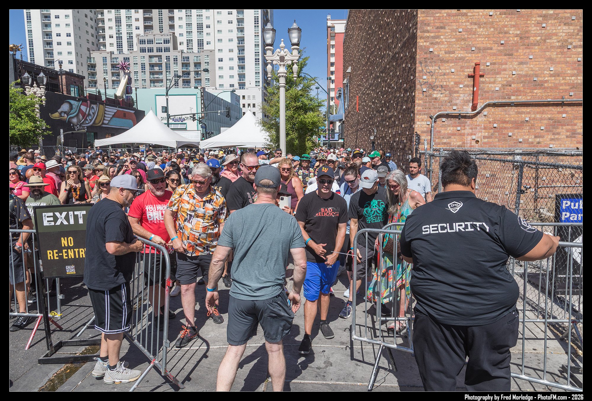 Crowd of people gathered behind a security barricade outside on a sunny day, with city buildings in the background.