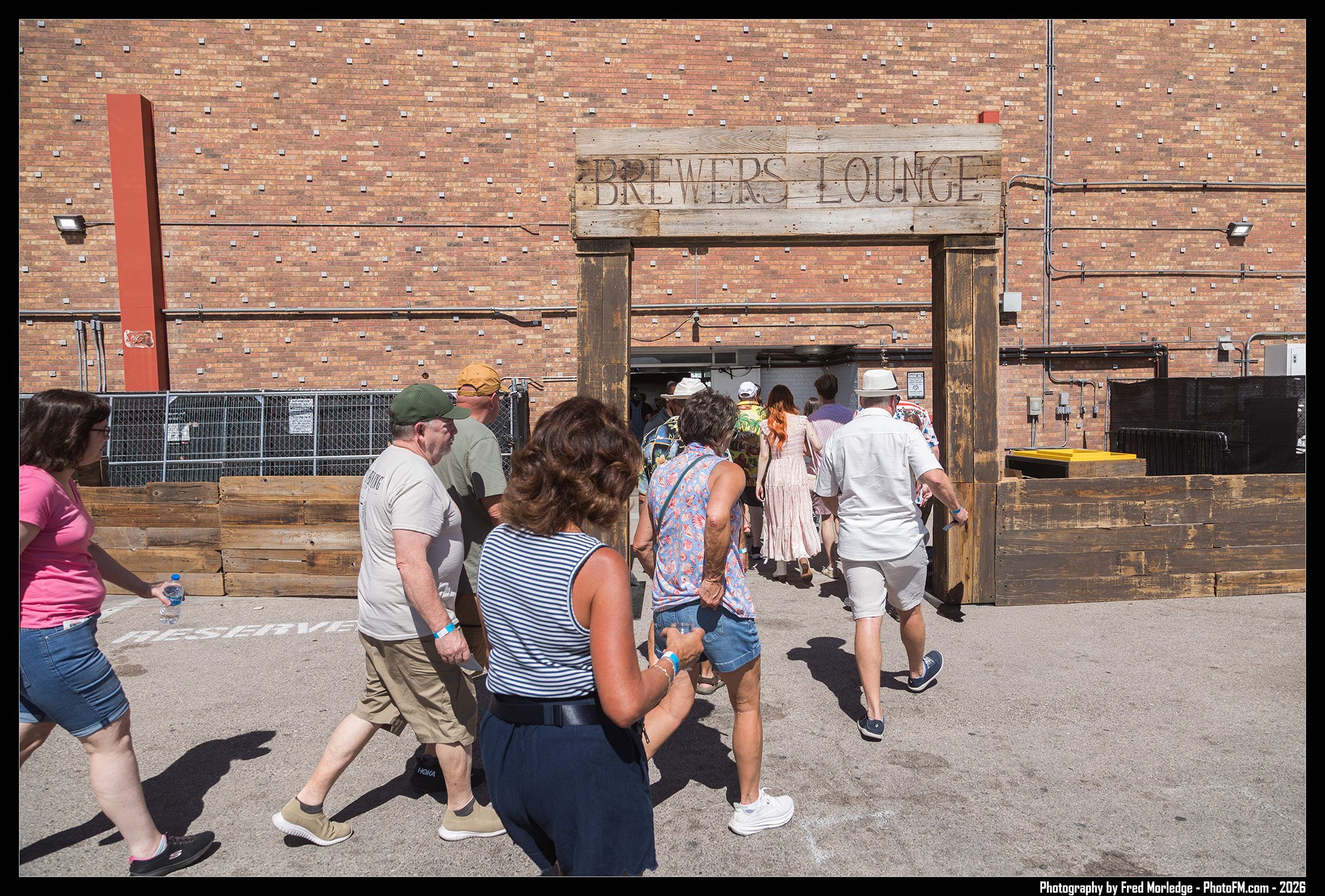 A group of people walking in line outside an entrance to a brewery lounge, with a brick wall and wooden sign overhead that reads 'BREWERS LOUNGE'.