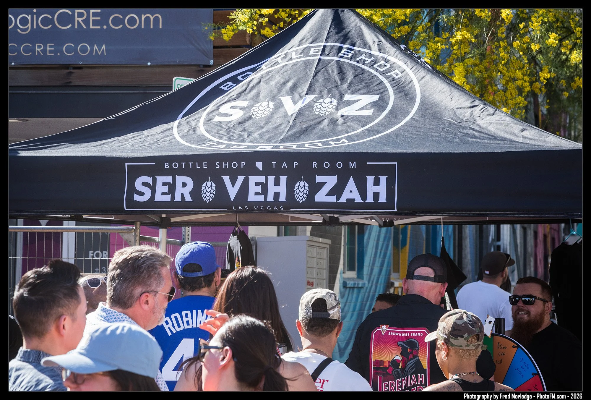 Crowd of people gathered at an outdoor event beneath a black canopy with white text reading 'serVEHZ' and 'bottle shop tap room,' with a yellow flowering tree and other banners in the background.