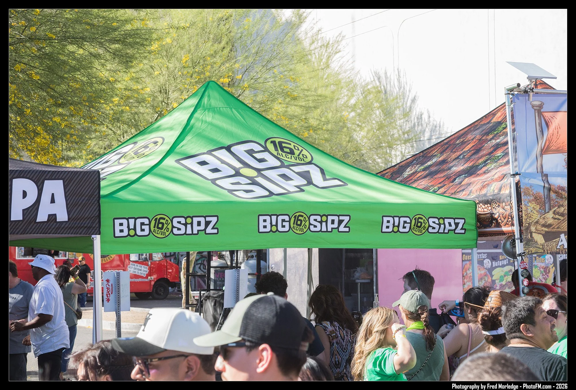 A group of people gathered under a bright green tent with the text 'BIG SIPZ' and '16% Alc/Vol' printed on it, at an outdoor event with trees and vendors visible in the background.