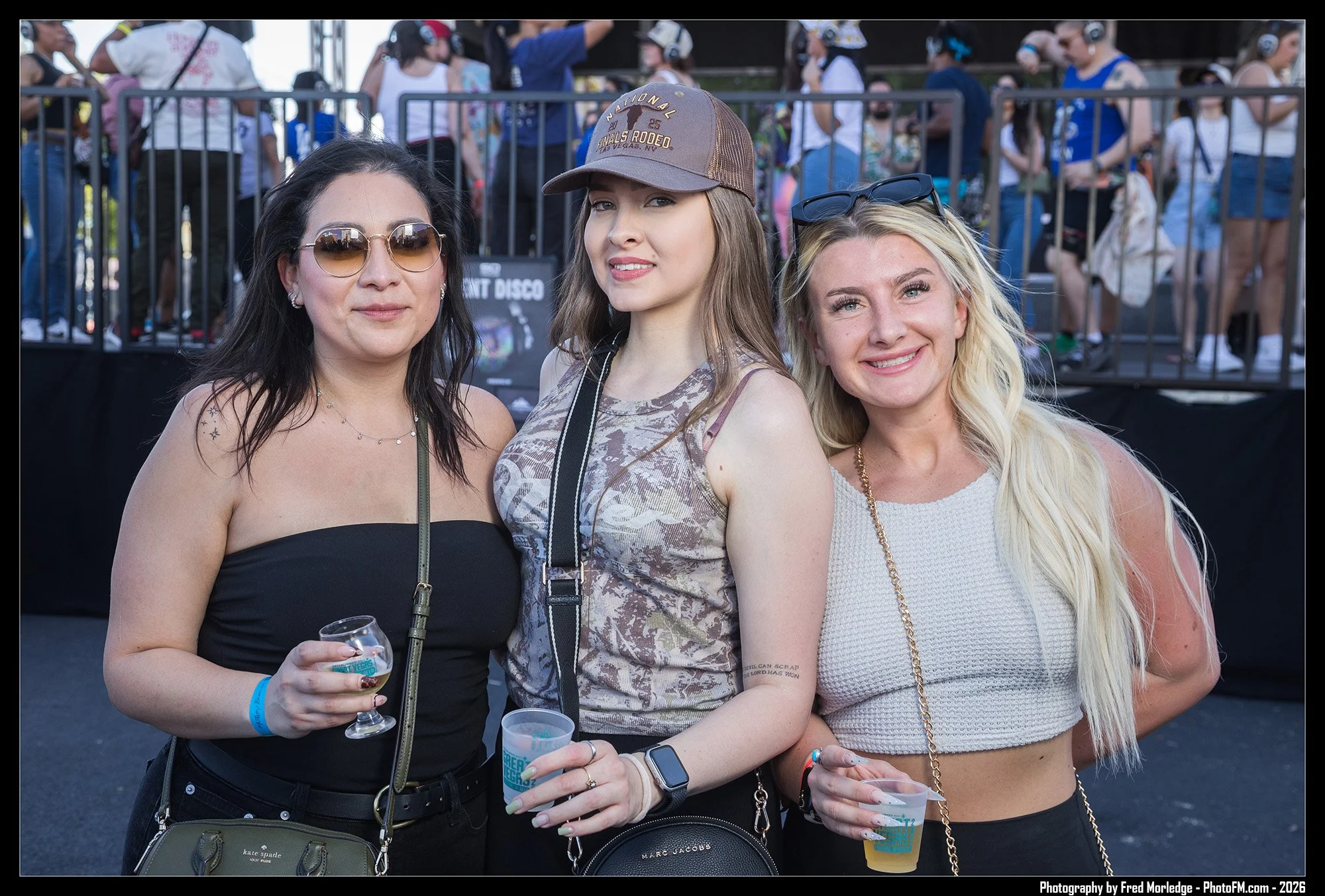 Three women posing at an outdoor event, with one holding a drink in her hand, all smiling at the camera. The woman on the left has dark hair, sunglasses, and a black top. The woman in the middle has light brown hair, a brown cap, and a patterned top.