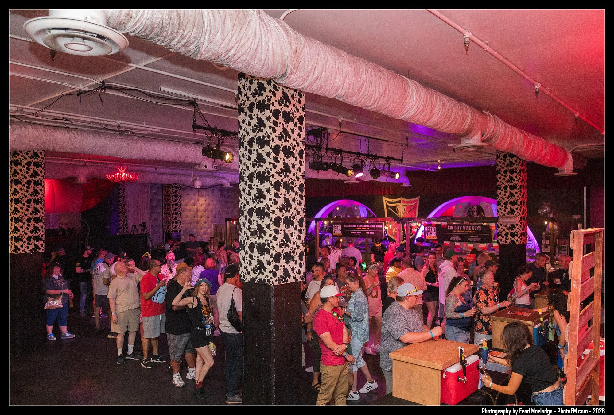 Crowd of people gathered indoors with some standing in line, with colorful lighting, patterned black and white columns, and a DJ booth at the back.