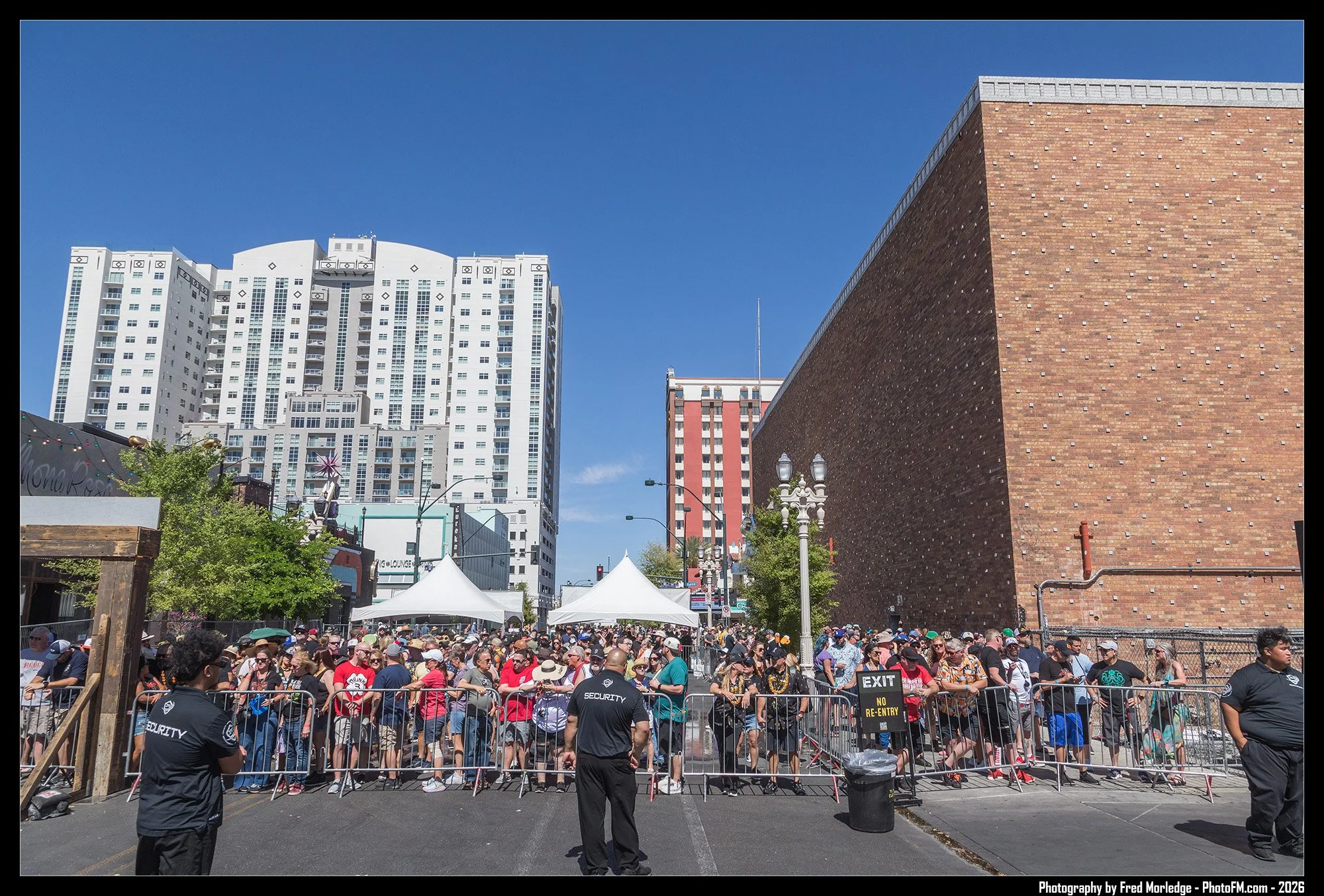 A large crowd of people gathered behind barricades at an outdoor event, with security personnel in the foreground. Tents and signs are visible in the background, along with tall buildings under a clear blue sky.