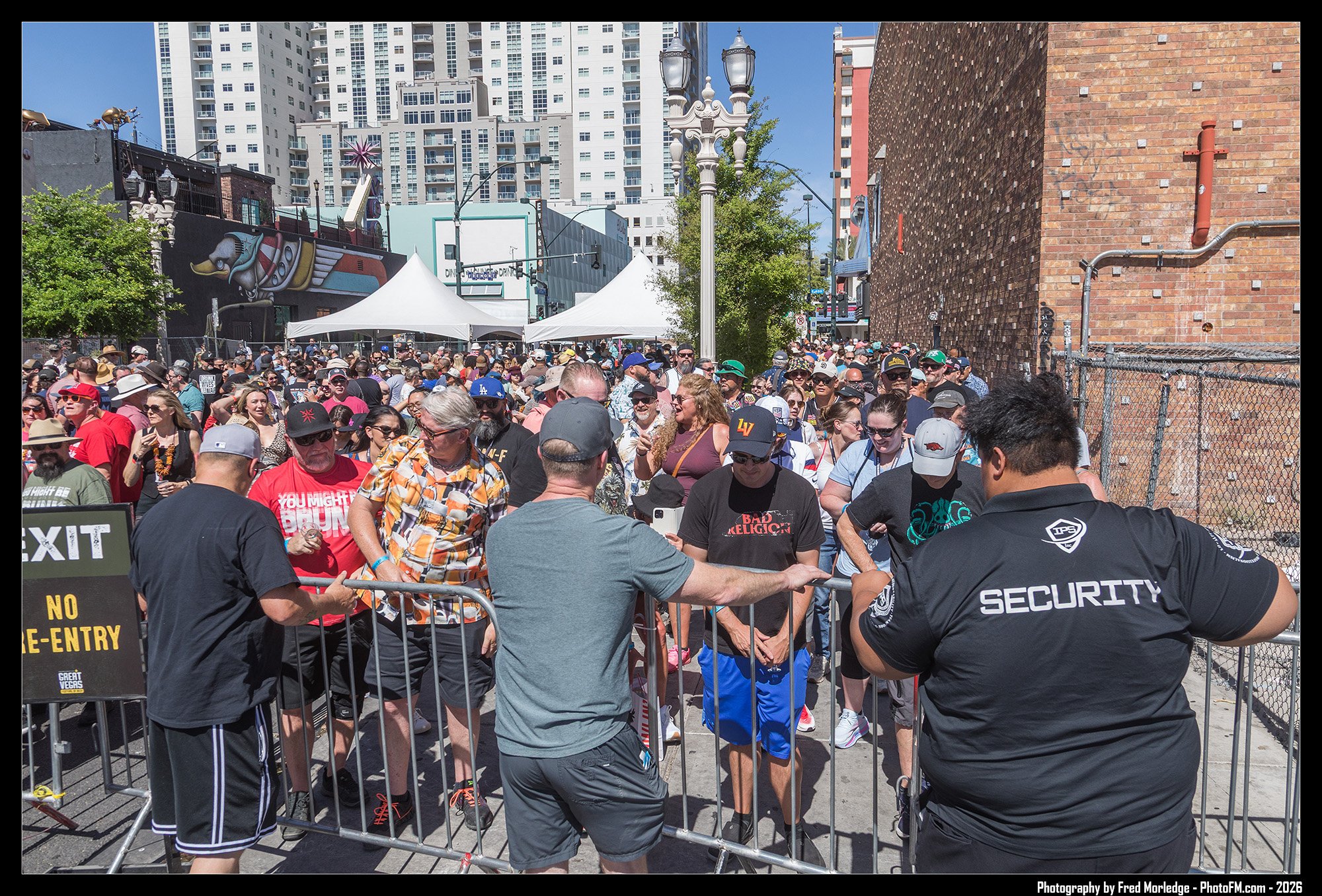 Crowd of people waiting in line outside an event entrance, with security guards and barriers, in an urban area with tall buildings and street art on a building in the background.