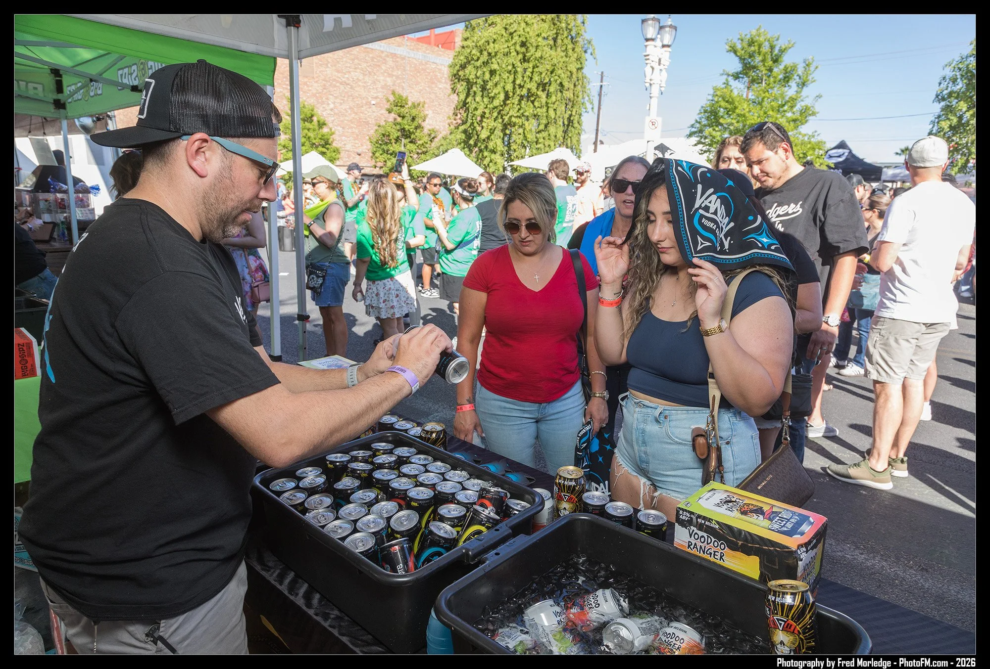 A man with glasses and a black hat sells cans of Voodoo Ranger beer to a group of women at an outdoor event. The women are gathered around his booth, some wearing sunglasses and one with a bandana on her head. The background shows crowd, tents, and t