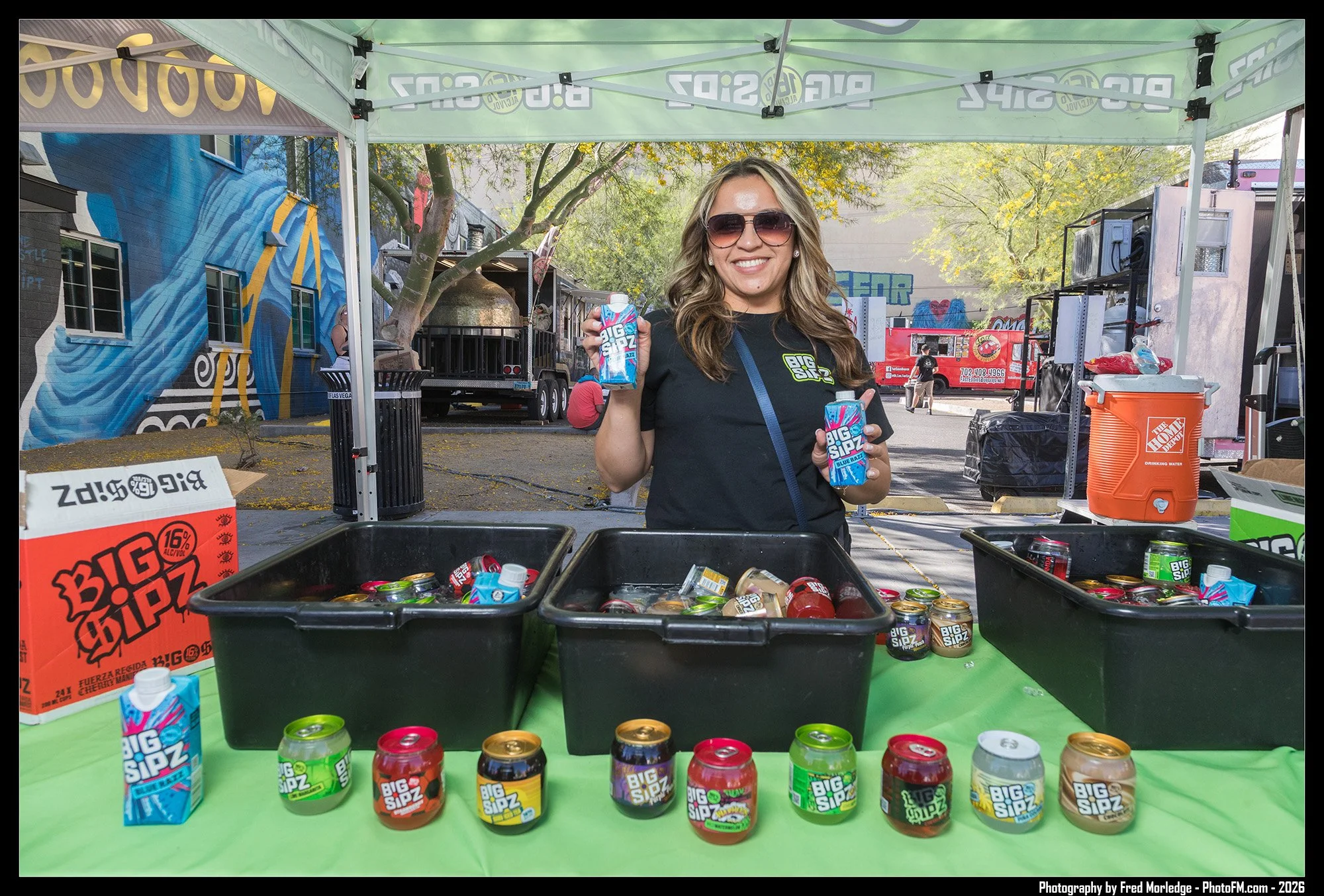 A woman smiling and holding cans of Big Sipz energy drink at an outdoor event, standing behind a table with several more cans of Big Sipz and a bin filled with cans, under a green pop-up tent with a mural wall in the background.