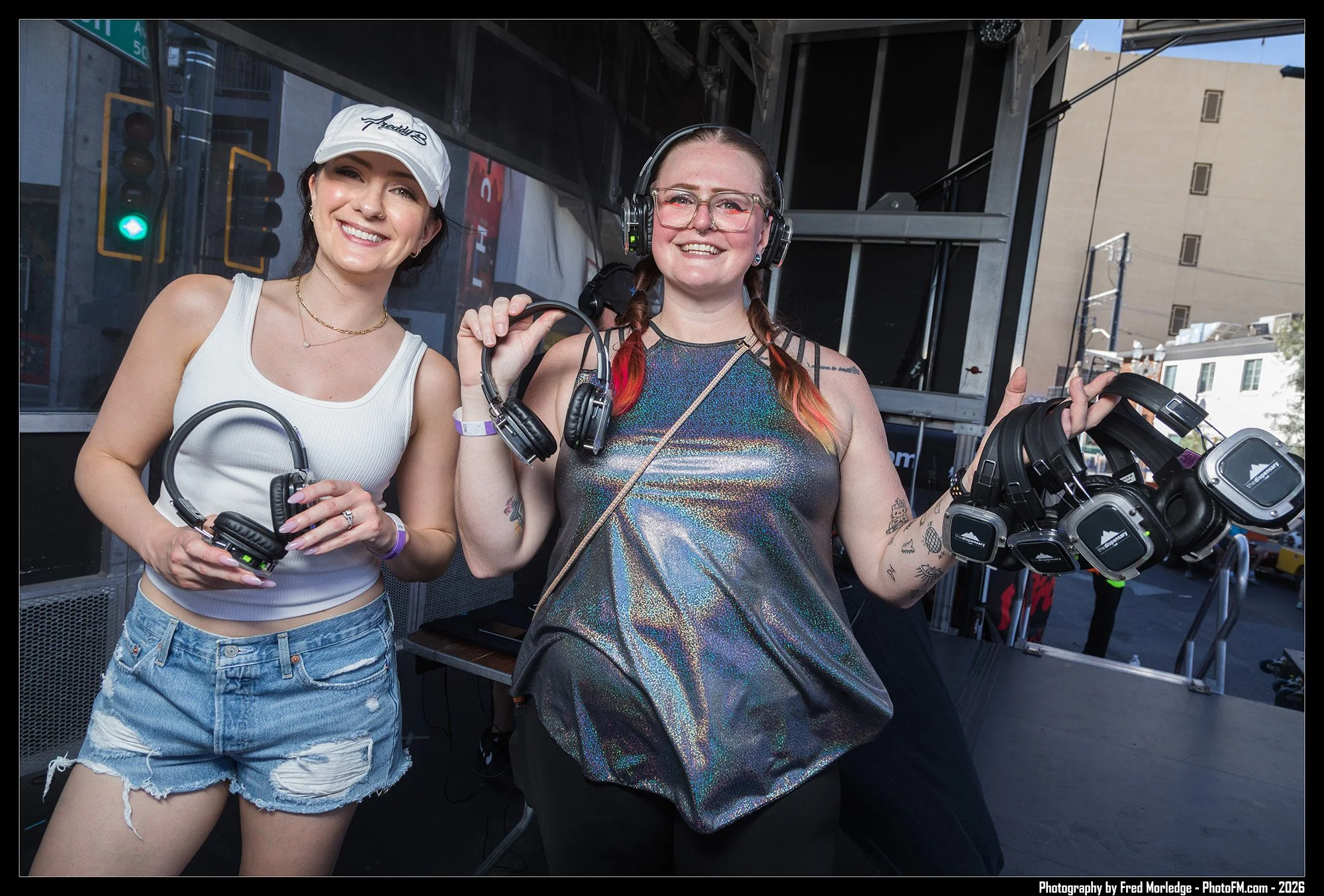 Two women smiling and holding headphones at an outdoor event, with a metal stage structure behind them and city buildings in the background.