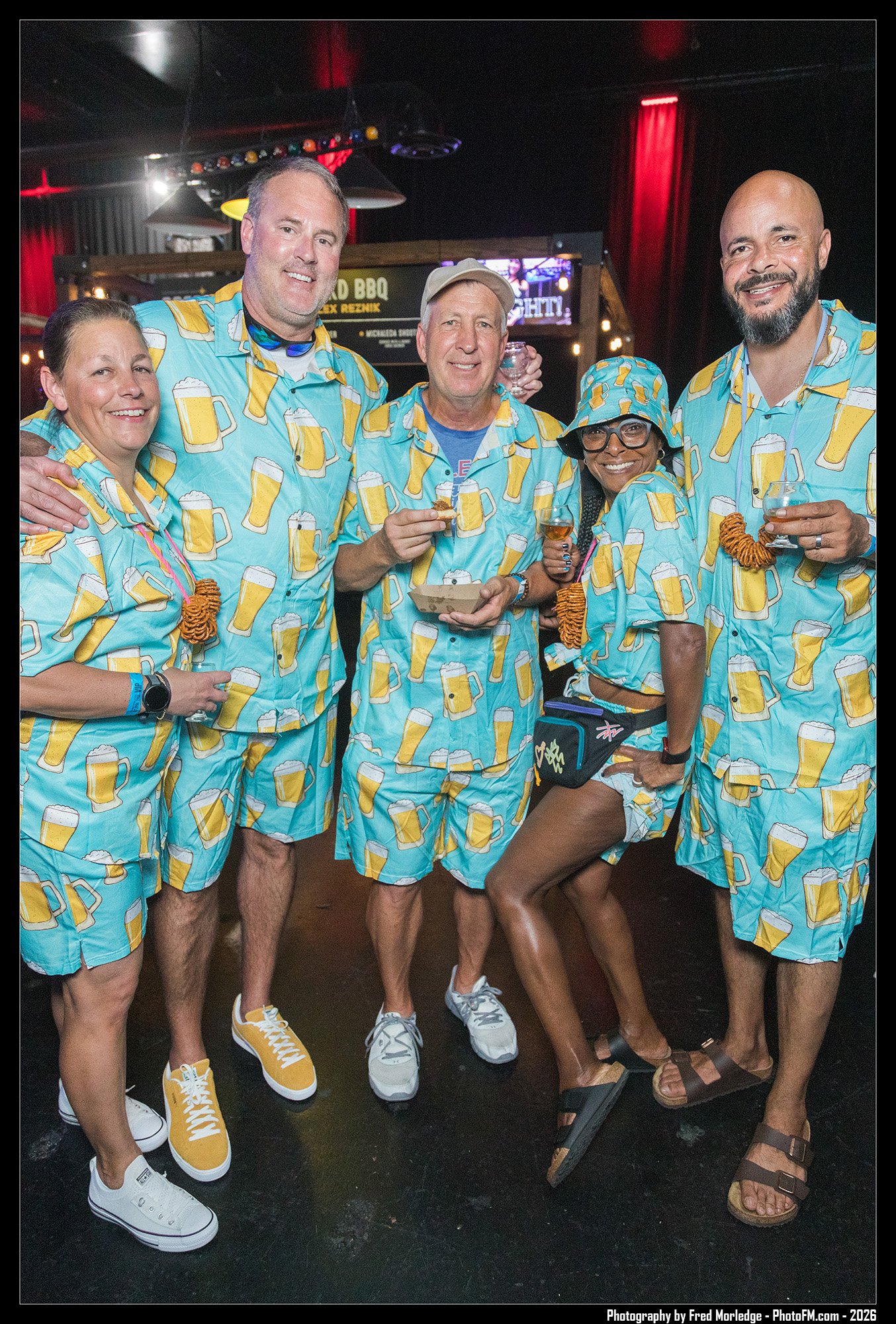Group of six people having fun at a social gathering, all wearing matching beer-themed shirts with beer mug patterns, in a dimly lit indoor venue with red curtains and a sign in the background.