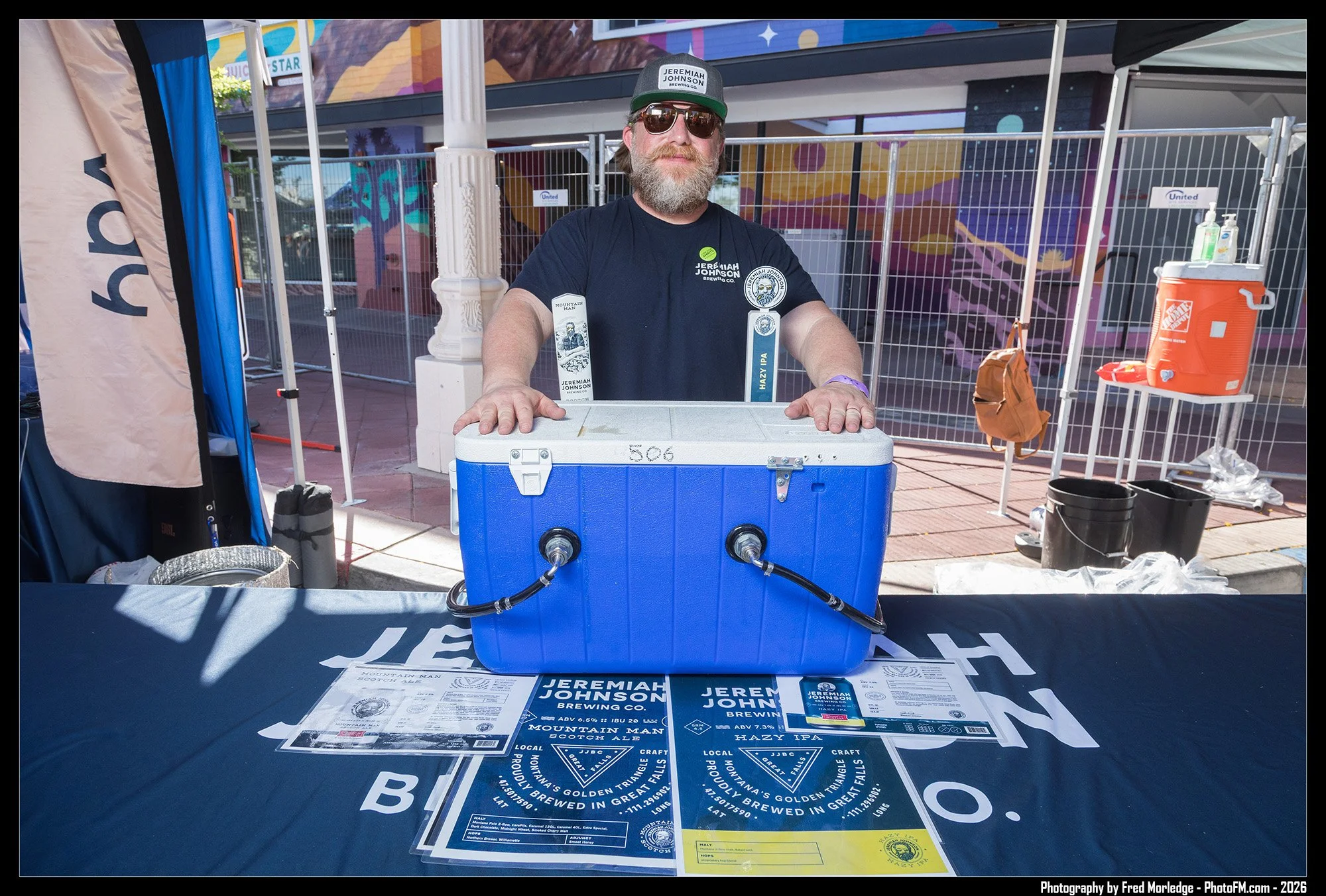A bearded man with sunglasses, a jacket, and a cap standing behind a table with beer tasting flyers, at an outdoor event. Behind him is a mural and fencing, with various event equipment and supplies nearby.