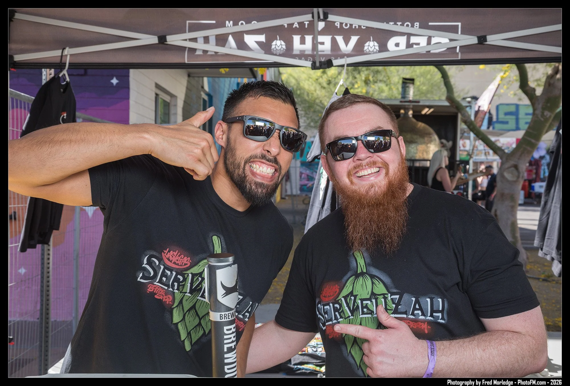 Two men with beards and sunglasses smiling at a booth at an outdoor event, wearing black t-shirts with green and red graphic designs. One man is pointing at the other. There is a canopy overhead and trees in the background.