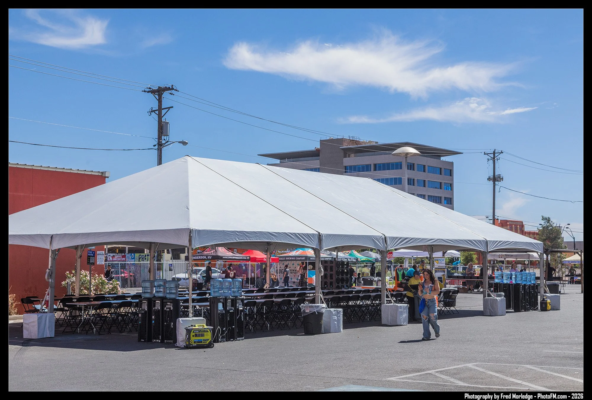 Outdoor event in a parking lot with multiple white tents, tables, and chairs. People are walking and sitting, with vendor booths visible under the tents. There are water bottles on the tables and a portable generator on the ground. A large building a