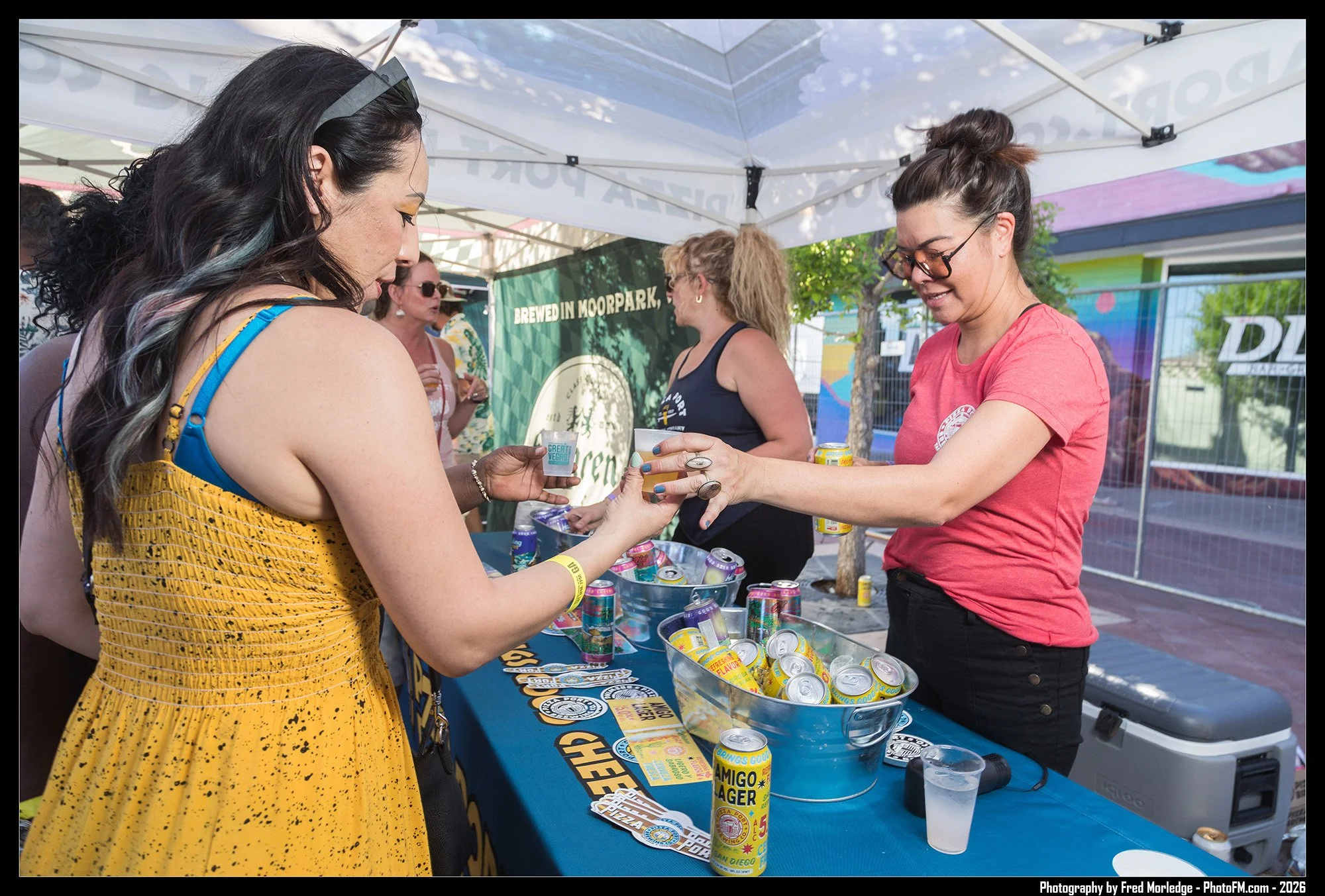 People enjoying a beverage at an outdoor event with a booth selling canned drinks. A woman in a yellow dress is reaching out to the woman behind the booth, who is wearing glasses and a red shirt. The booth has cans of drinks on the table and a sign t
