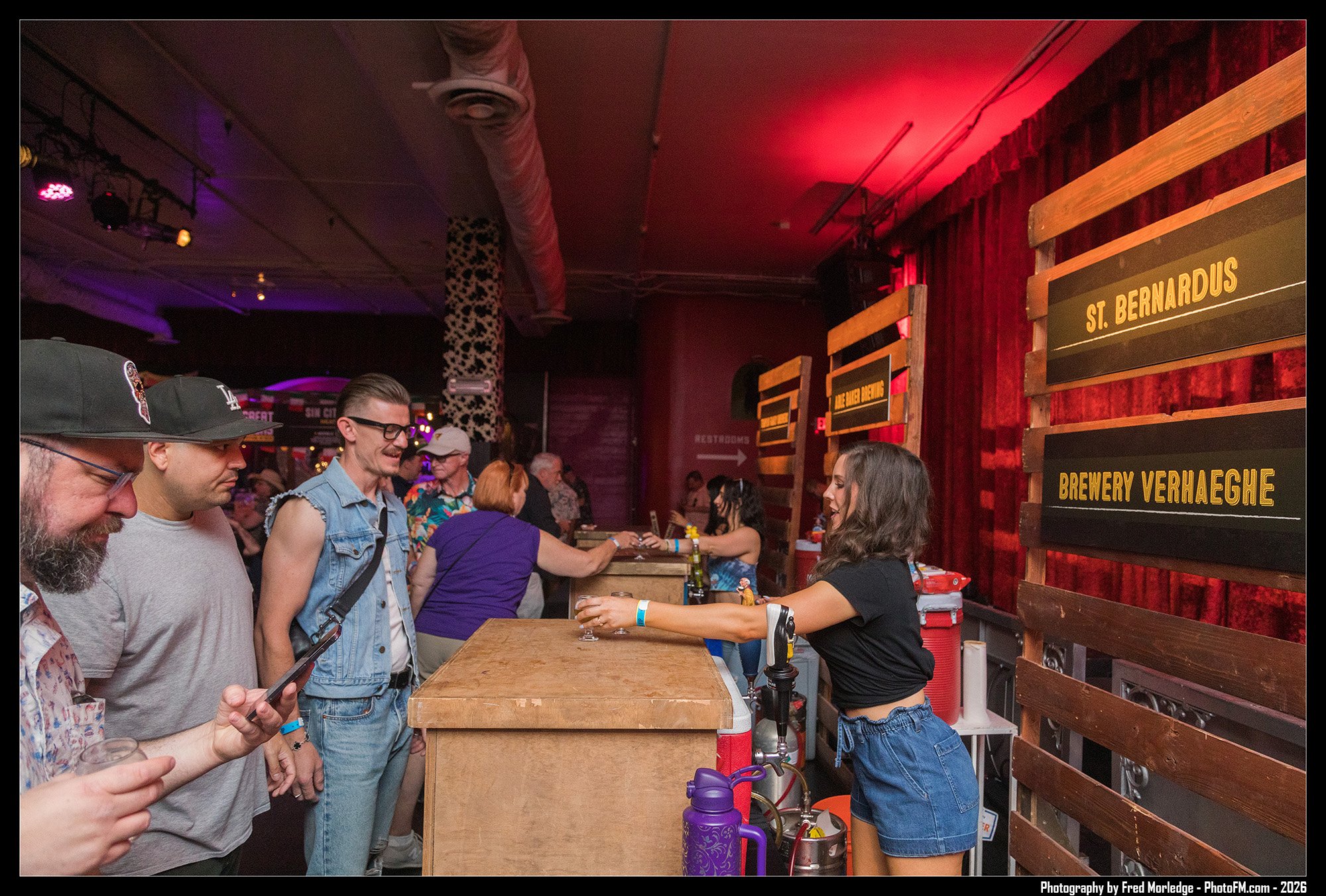 People standing in line at a bar with wooden signage reading 'St. Bernardus' and 'Brewery Verhaeghe' in a dimly lit venue with red lighting and decorative wall patterns.