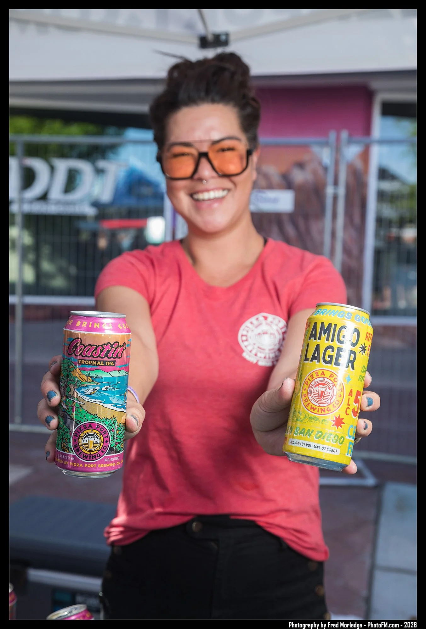 A smiling woman with short dark hair, glasses, and a septum piercing, wearing a pink t-shirt, holding two colorful cans of craft beer toward the camera, outdoors in front of a background with a sign and a fence.