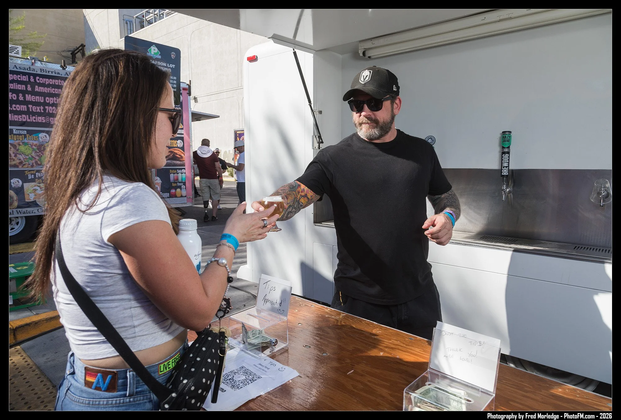 A woman with long brown hair wearing sunglasses and a white crop top is at a food truck, receiving a drink from a bearded man wearing a black baseball cap, sunglasses, and a black t-shirt. The food truck has taps for beer or other beverages, and ther