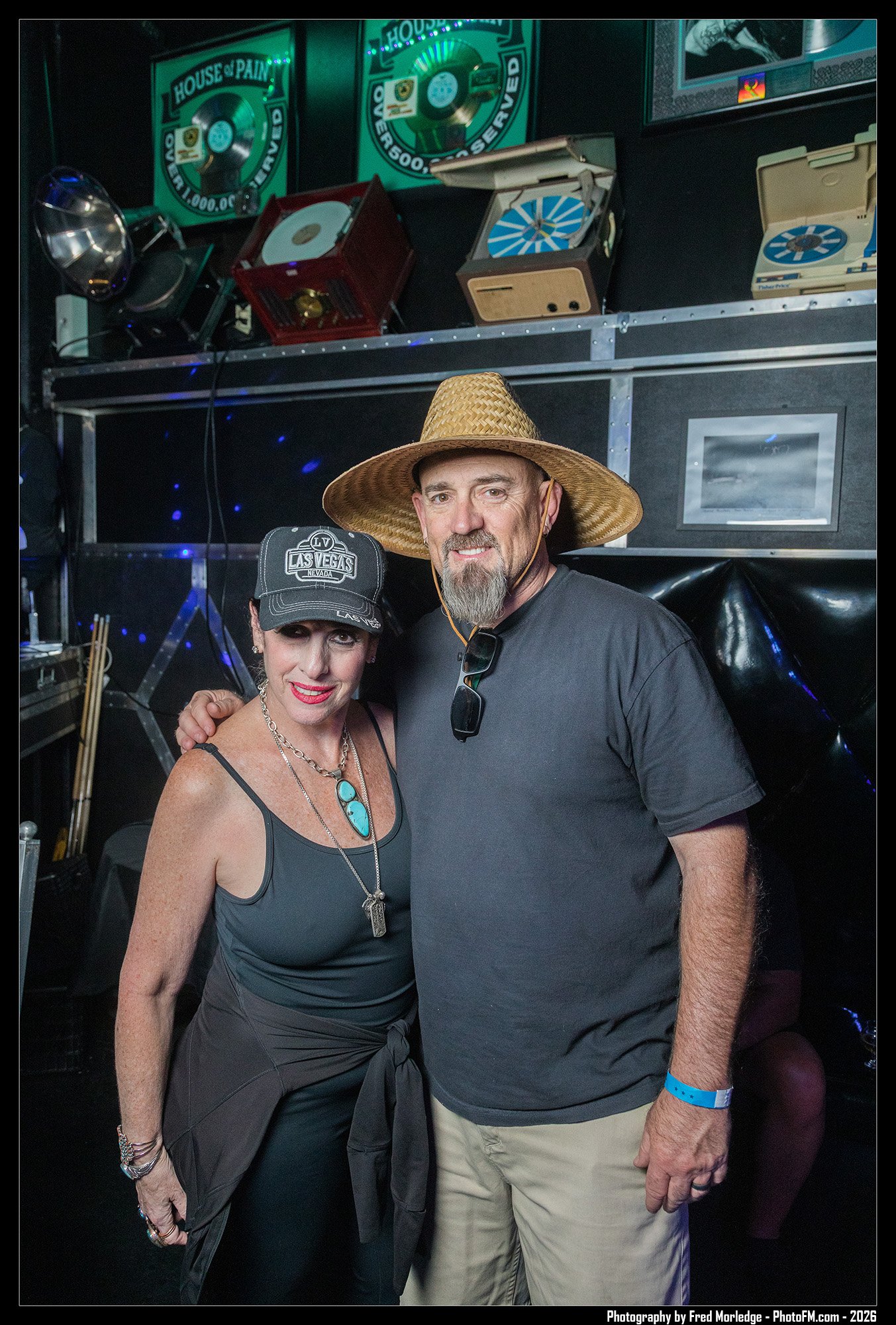 Two people, a woman wearing a black cap, black tank top, and jewelry, and a man in a gray t-shirt with a large straw hat, posing together indoors, with music and memorabilia in the background.