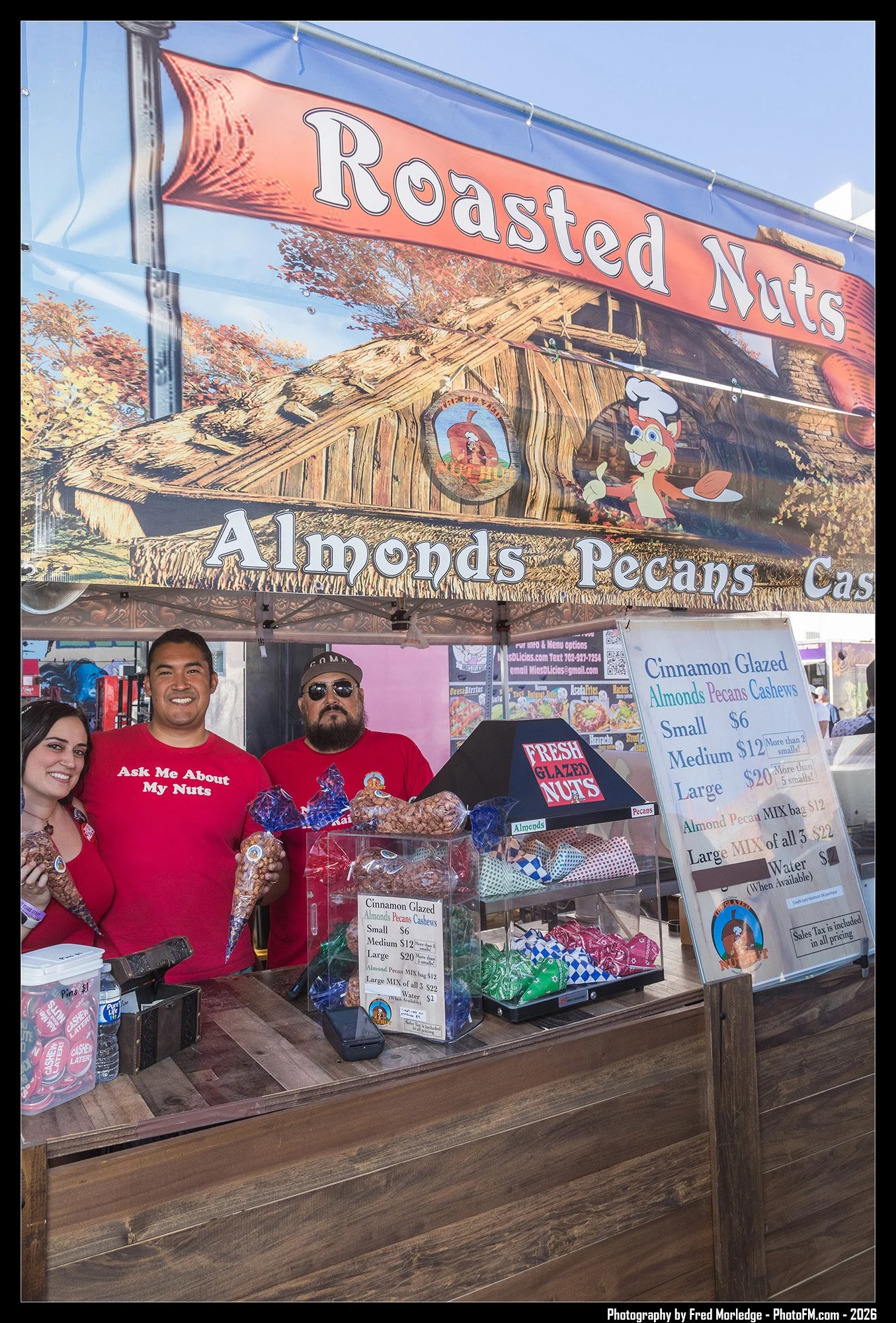 A fair booth named 'Roasted Nuts' offers candied almonds, pecans, and cashews in small, medium, and large sizes. Three vendors, two men and a woman, stand behind the booth smiling, one man has a beard and sunglasses, and they wear red shirts. A sign 