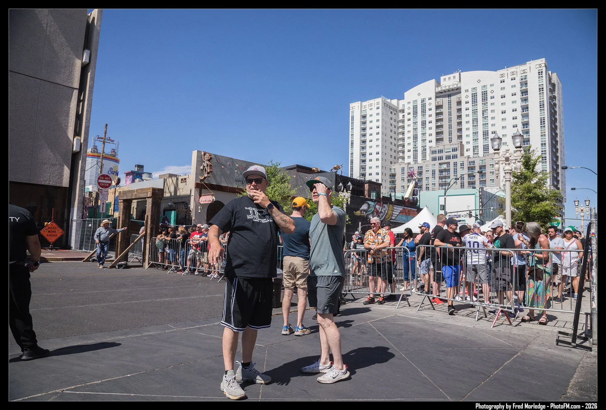 People waiting in line behind metal barricades on a city street, with some talking and others looking in different directions, with tall buildings and a clear blue sky in the background.