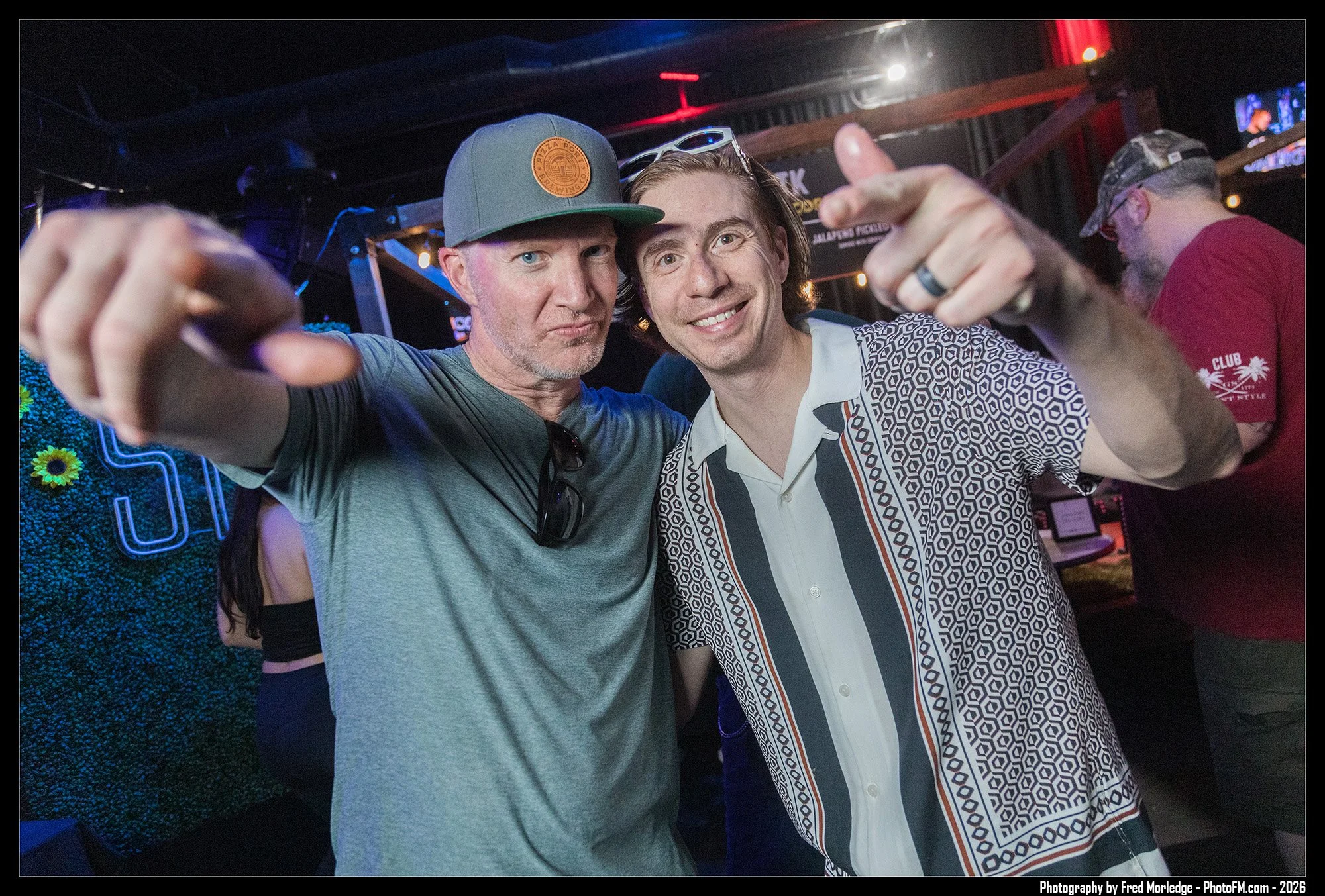 Two men posing closely together, one wearing a gray baseball cap and the other in a patterned shirt with sunglasses on his head, in a lively nightlife setting with colorful lights and other people in the background.