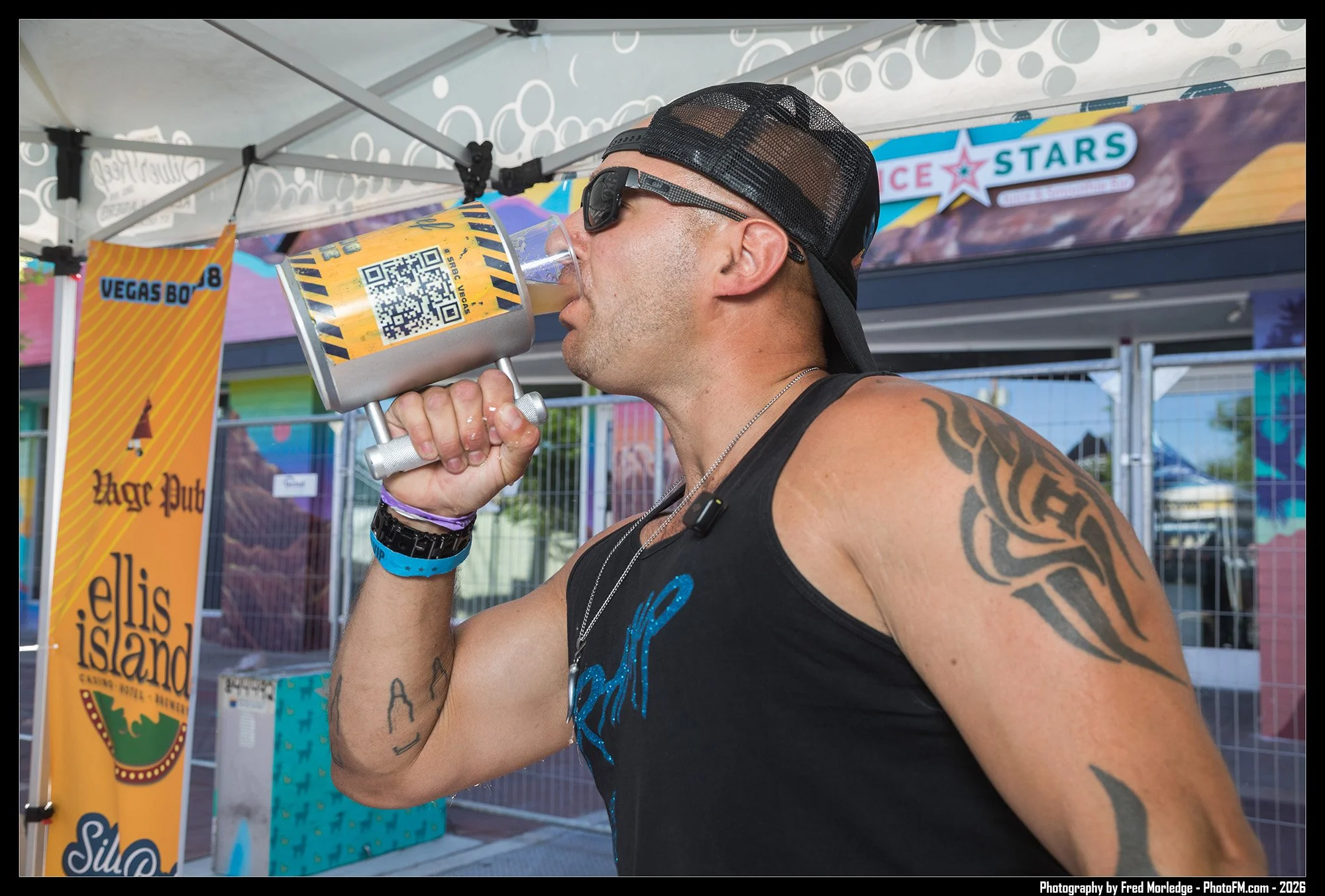 A man with tattoos on his arm wearing a black sleeveless shirt, sunglasses, and a black trucker cap, is drinking a beverage from a large container at an outdoor event. Behind him are colorful banners and signs, including one for Vegas 80s and a marqu