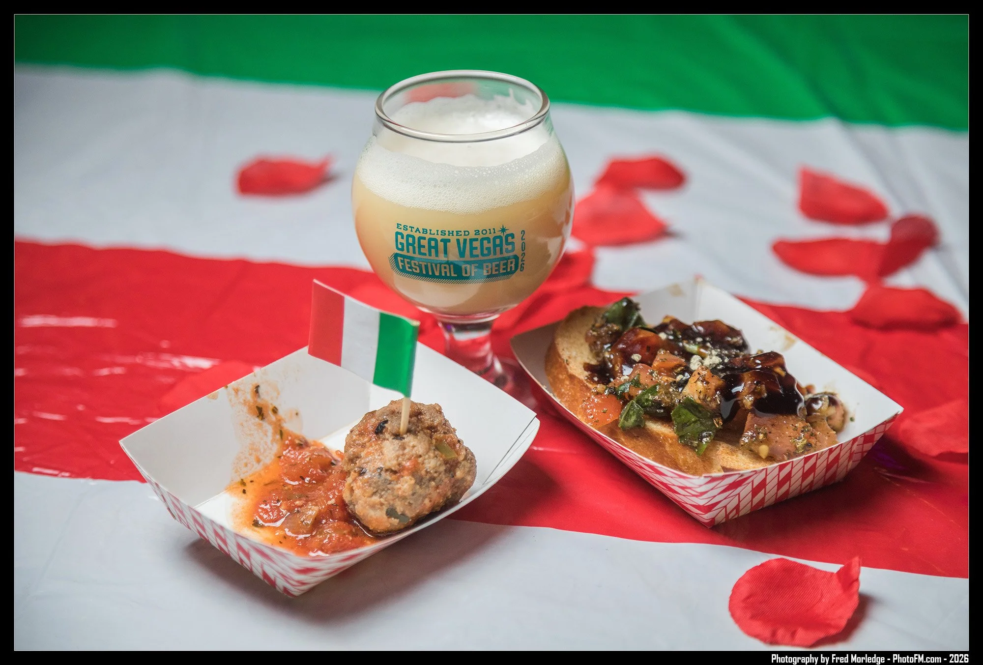 A glass with foam on top and text reading 'Great Vegas Festival of Beer' next to plates of food, including meatballs and bread with toppings, on a table with a red, white, and green tablecloth and red decorative petals.