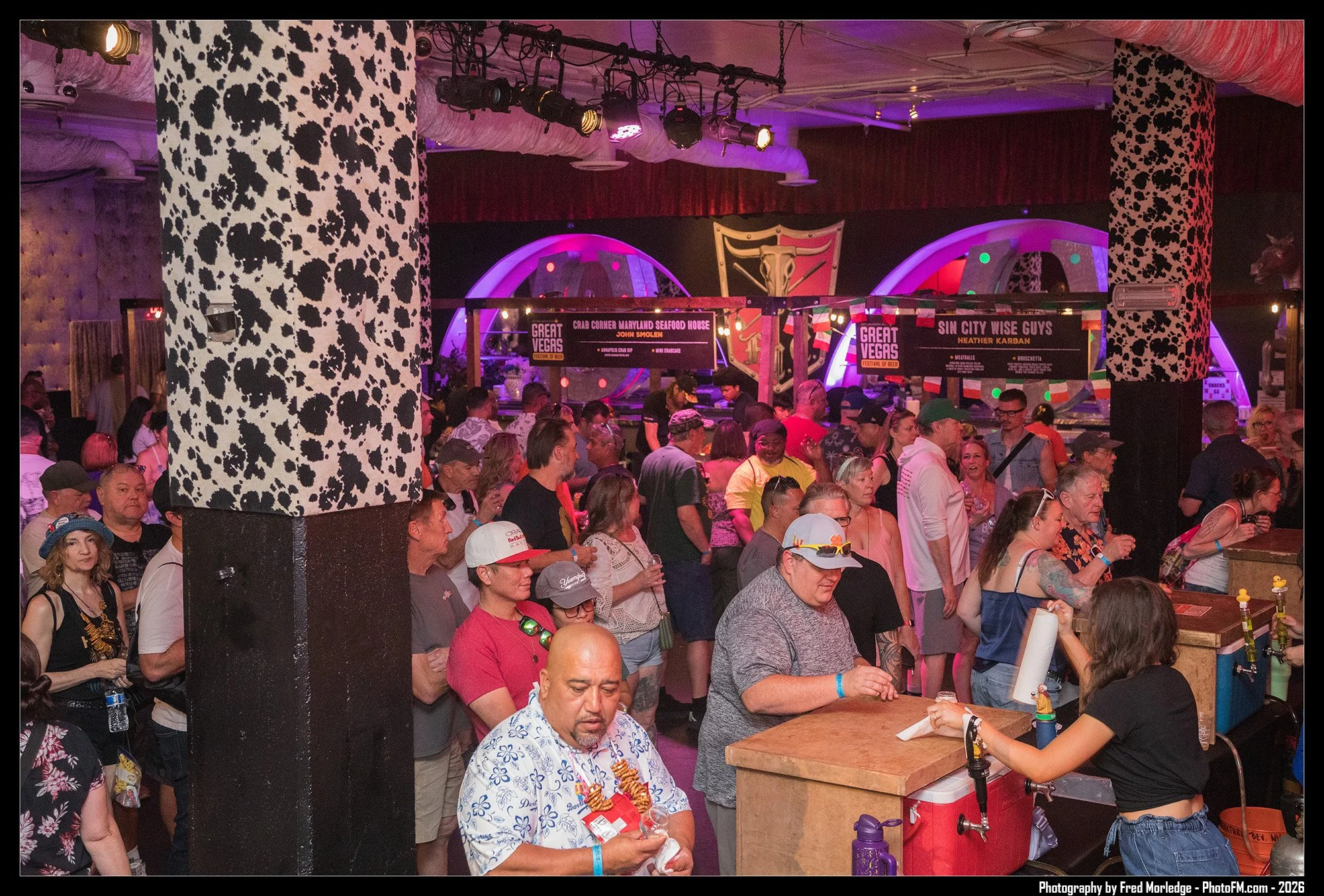 Crowd of people gathered indoors at a festival or event, with colorful stage lighting and signage for different food vendors in the background.