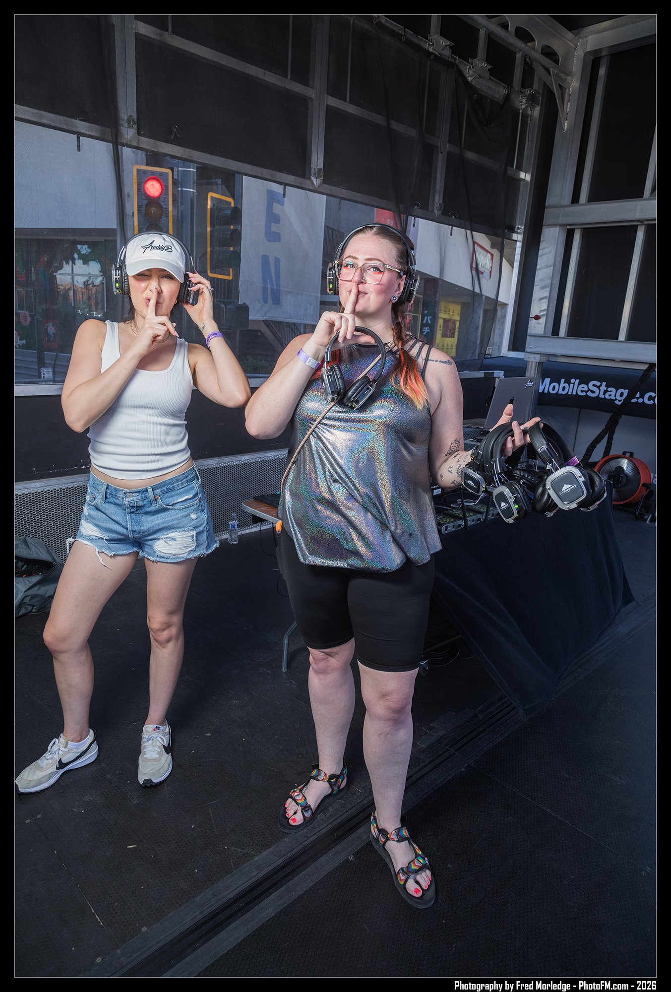 Two women on a stage, both wearing headphones and making 'shh' gestures with their fingers. The woman on the left wears a baseball cap and tank top, while the woman on the right wears glasses, a metallic top, and colorful sandals. There are DJ equipm