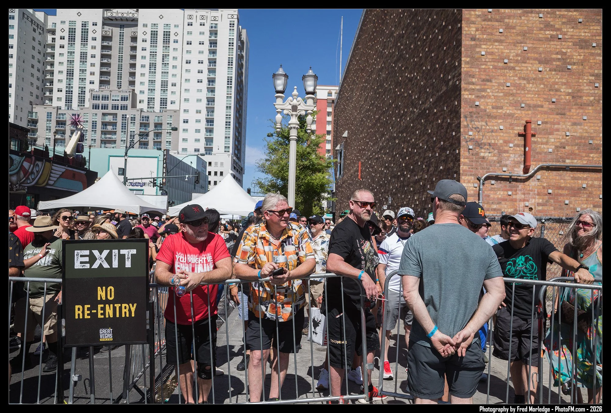 Crowd of people standing behind a barricade at an outdoor event with city buildings and a brick wall in the background. Some people are wearing sunglasses, hats, and casual clothing. A sign reads 'EXIT' and 'NO RE-ENTRY'.
