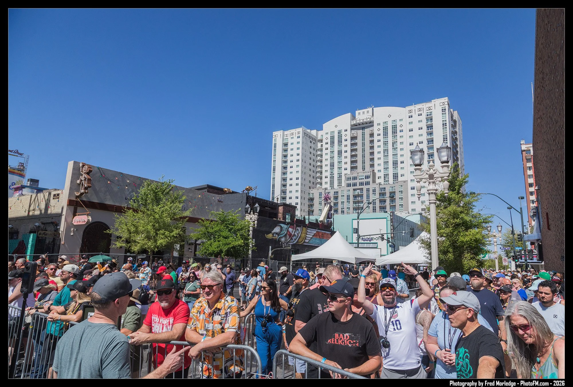 A large crowd of people gathered outdoors in front of a stage or event area, with a cityscape of tall buildings and blue sky in the background.