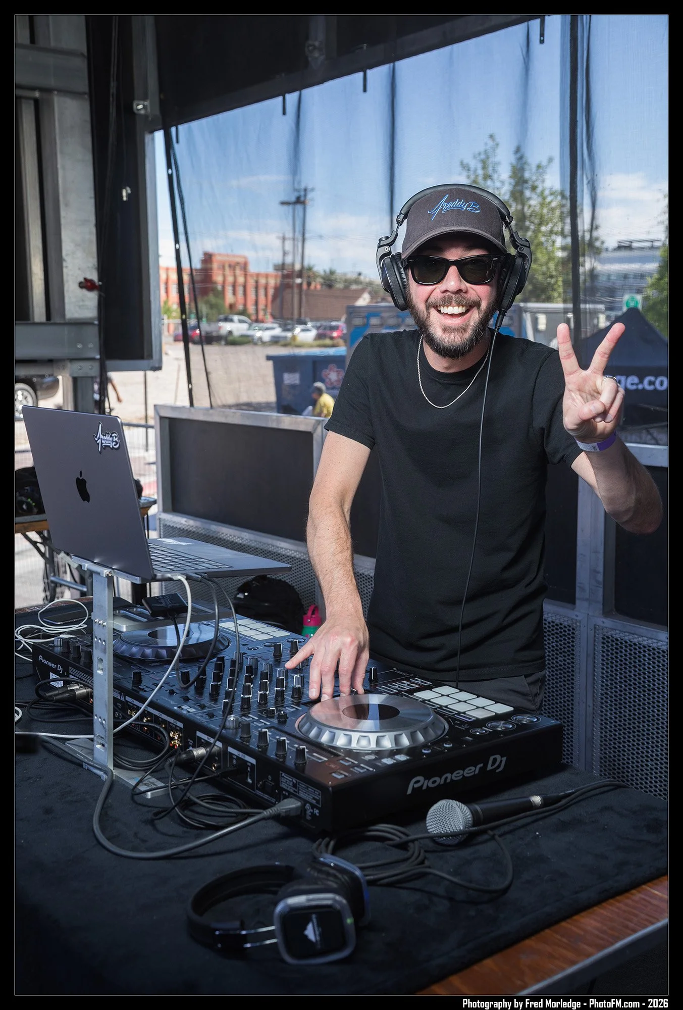 A man with a beard, sunglasses, and a black cap with blue writing is smiling and making a peace sign while DJing at an outdoor event. He is using a Pioneer DJ controller surrounded by headphones, a laptop, and other audio equipment.