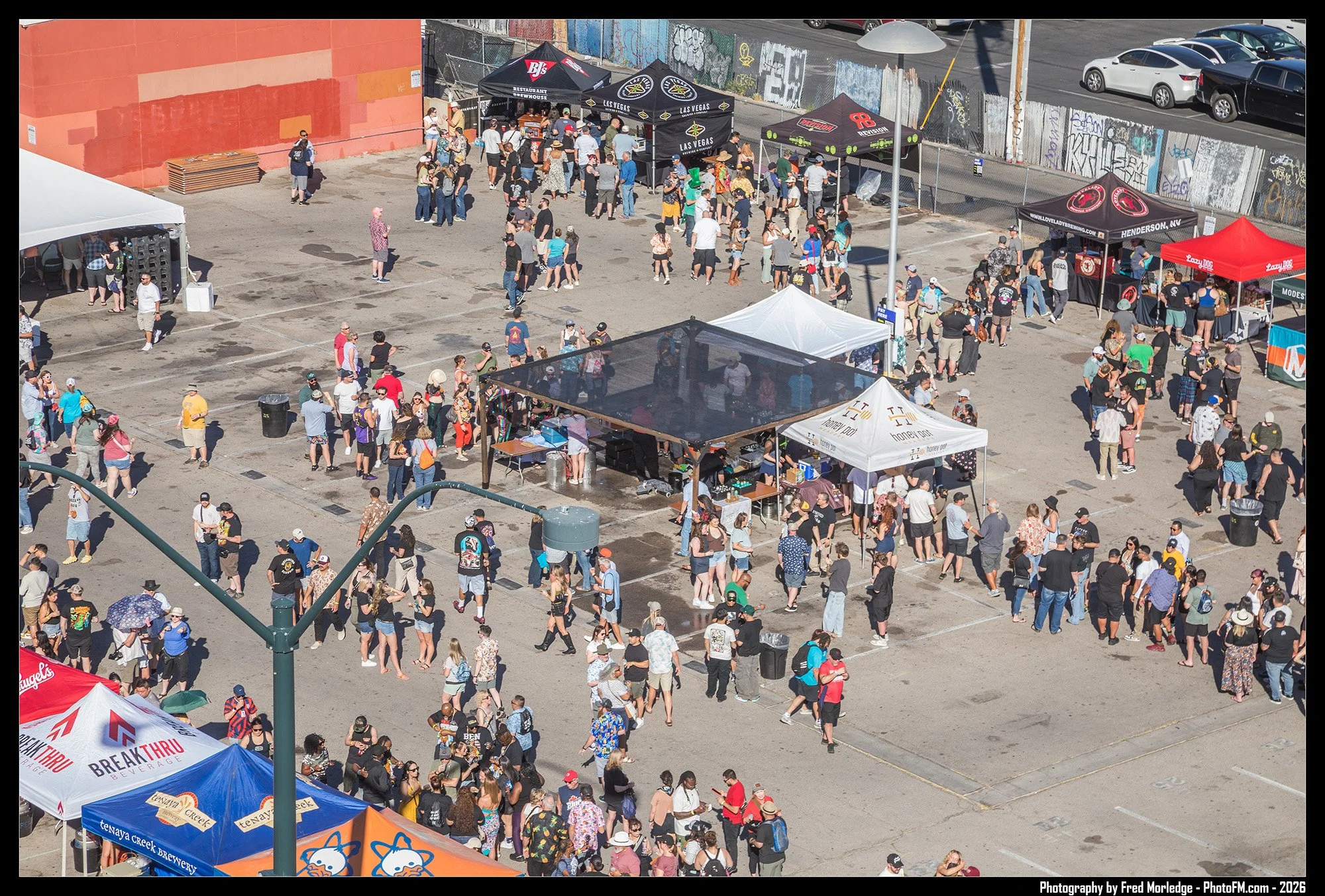 Crowd of people at an outdoor festival with vendor tents, some people are walking or standing, and there are traffic and graffiti on a fence in the background.