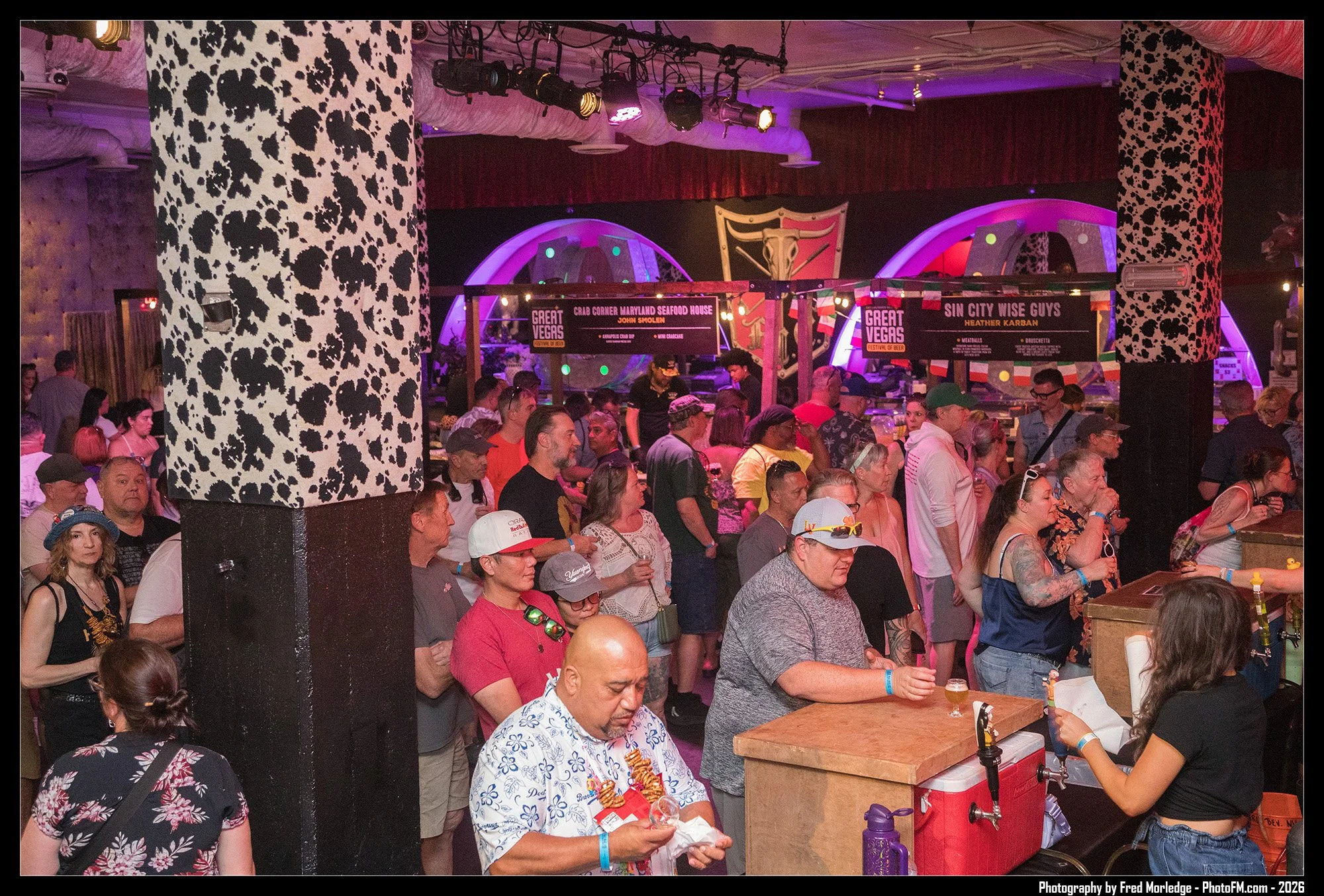A crowded indoor event with many people standing and sitting, colorful lighting, and signs indicating a Las Vegas festival of beer with food vendors.