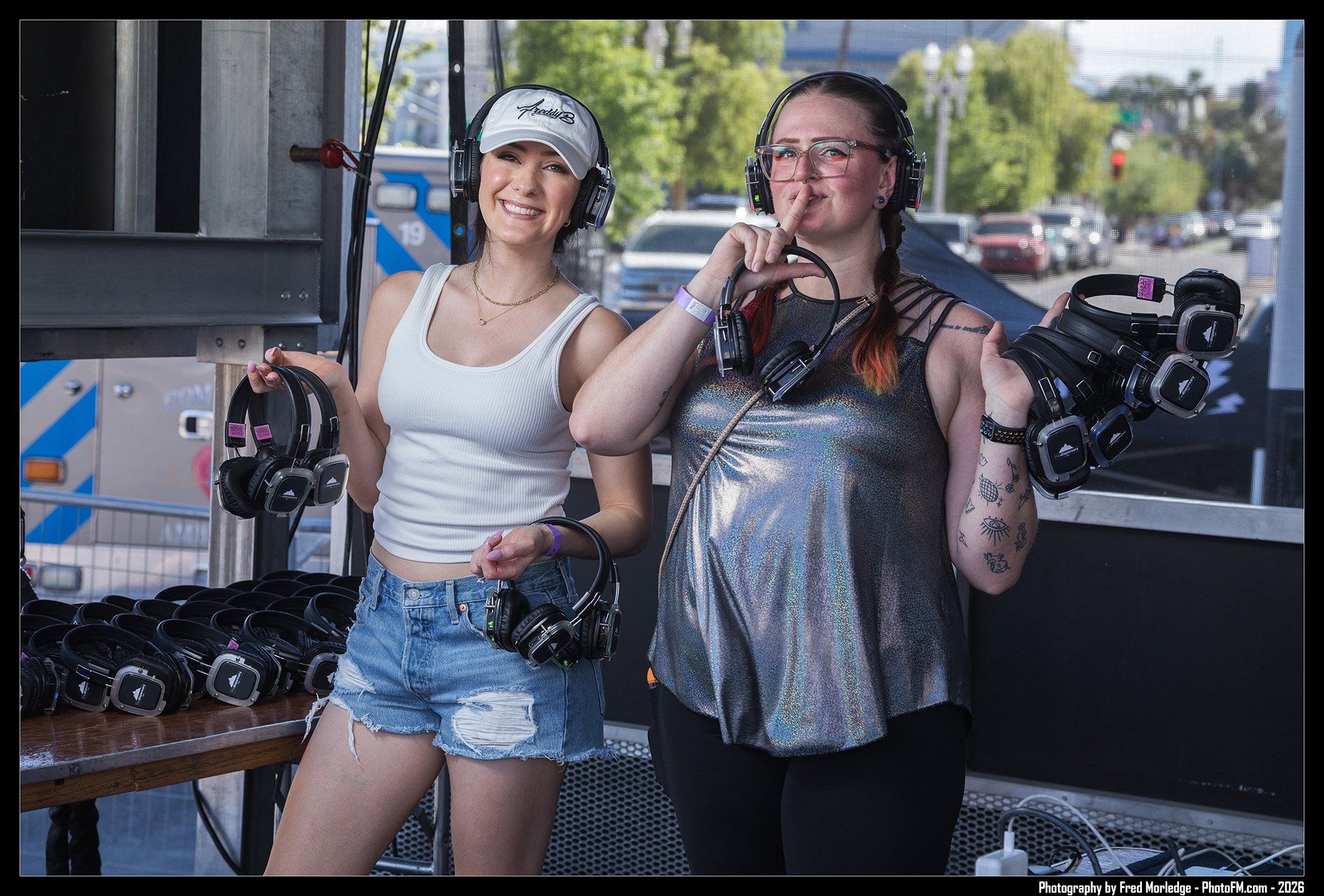 Two women standing behind a table of neatly arranged headphones at an outdoor event. The woman on the left wears a white tank top, denim shorts, a white cap, and headphones, smiling at the camera. The woman on the right carries headphones, wears glas