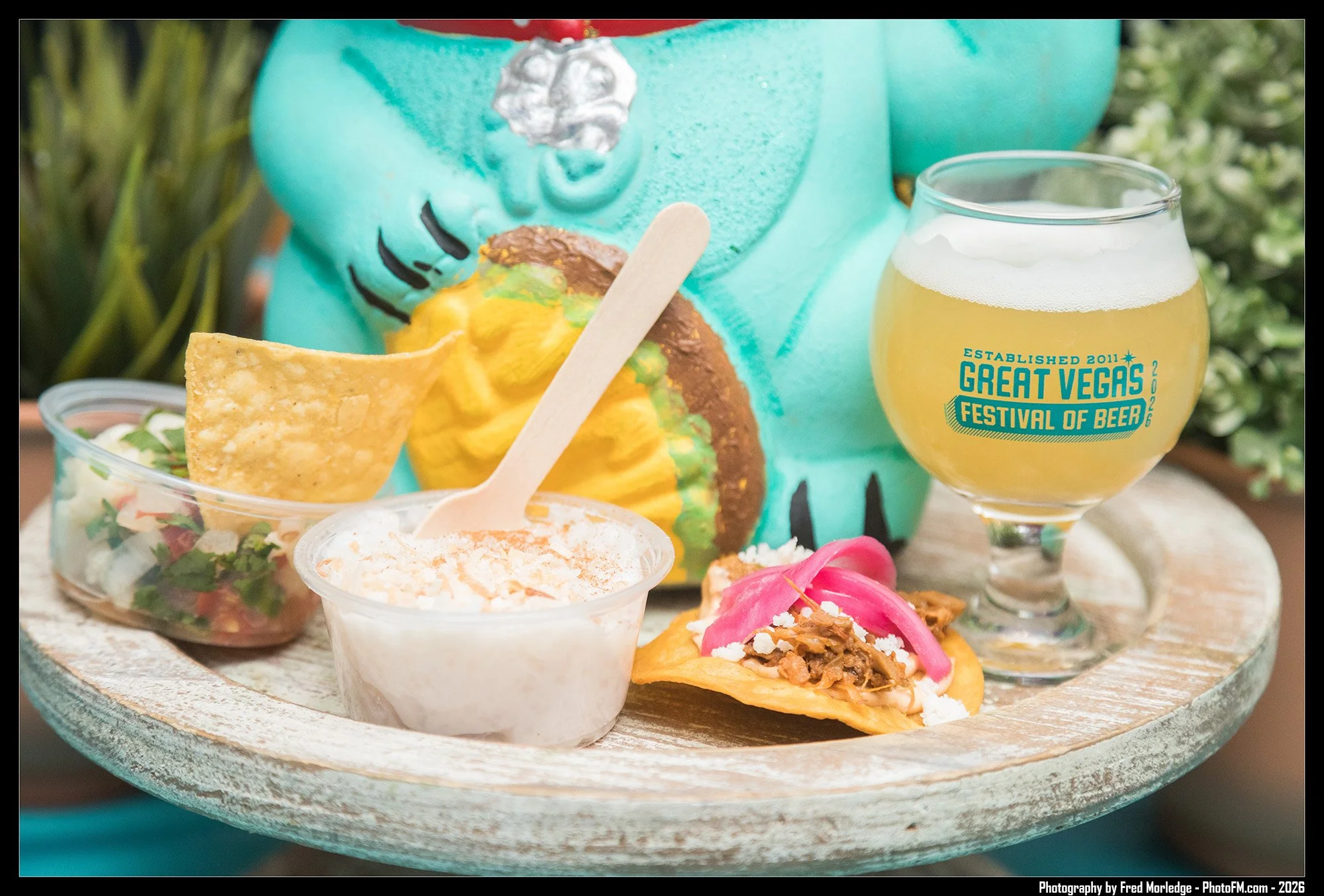A tray with various Mexican dishes including guacamole, nachos, tacos, and a small salad, along with a glass of beer in front, set against a colorful background.