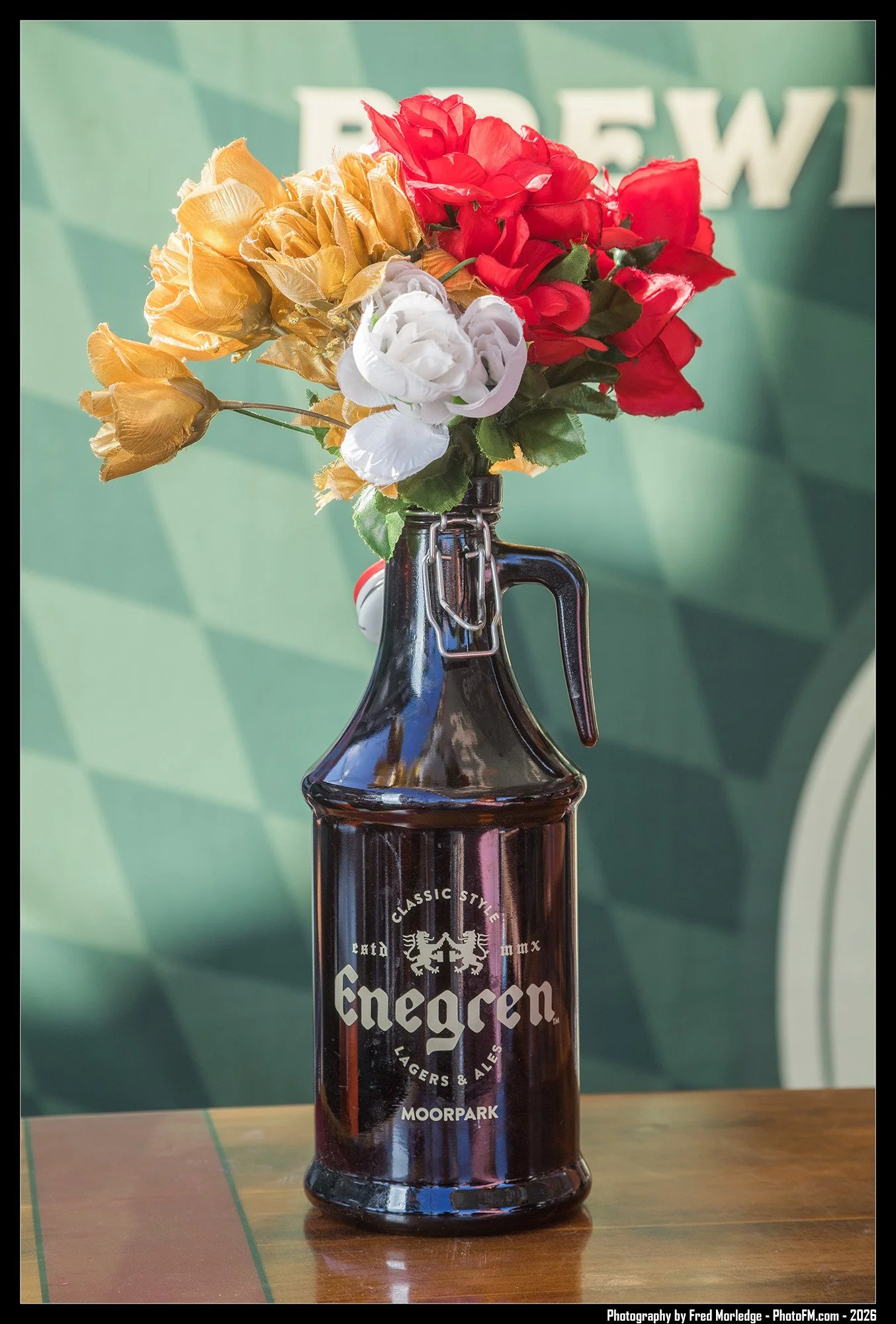 A glass bottle with a bouquet of artificial flowers including red, white, and yellow roses, placed on a wooden surface against a green background with part of a logo or sign showing the letters 'REW'.